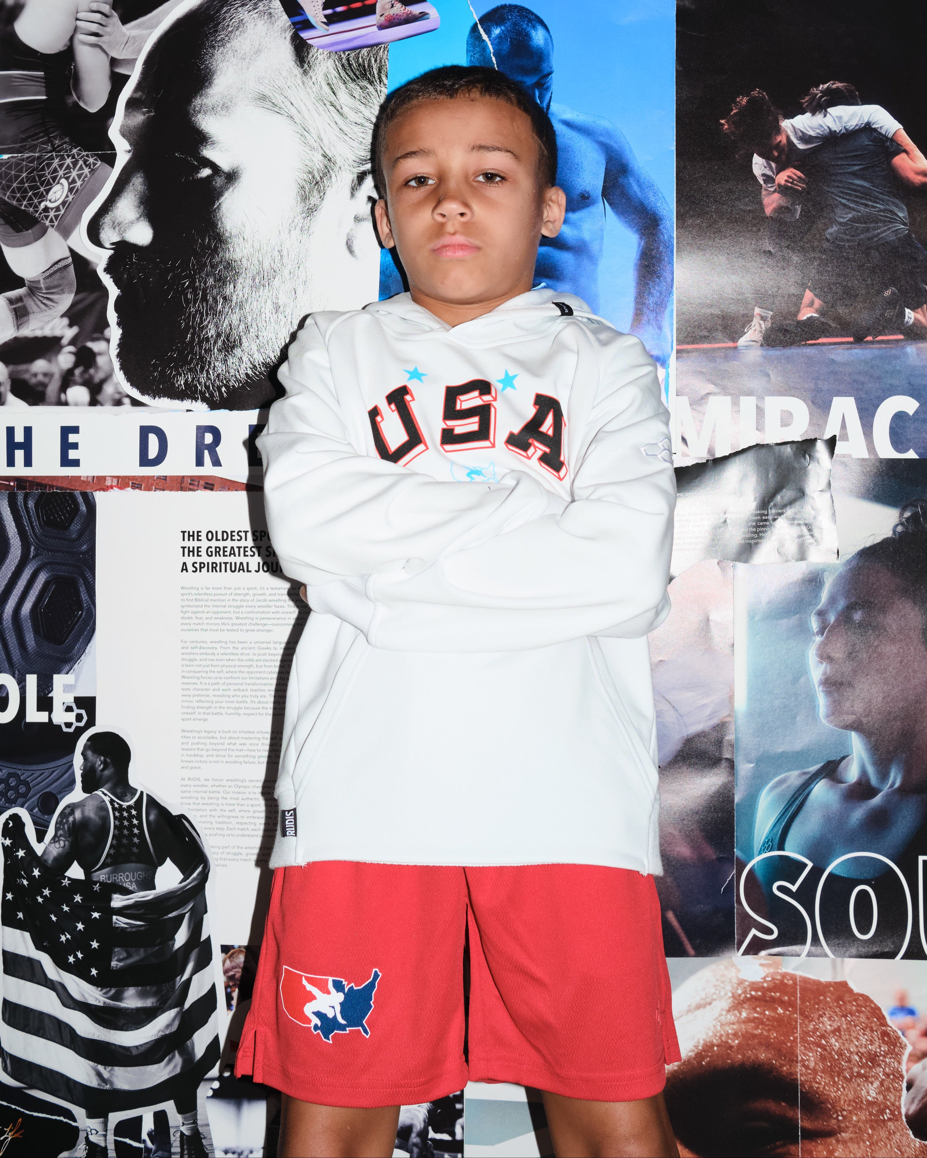 Beacon Burroughs, youth wrestler and son of Olympic champion Jordan Burroughs, wearing a white USA Wrestling hoodie and red USA Wrestling shorts by RUDIS, standing confidently in front of a wrestling mural — representing the next generation of wrestling.