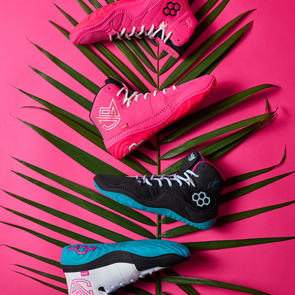 Editorial flat lay of the RUDIS JB1 South Beach Collection wrestling shoes, featuring the Flamingo Pink and South Beach colorways, styled against a tropical palm leaf and vibrant pink backdrop — highlighting the bold design and feature of the Jordan Burroughs South Beach collection.