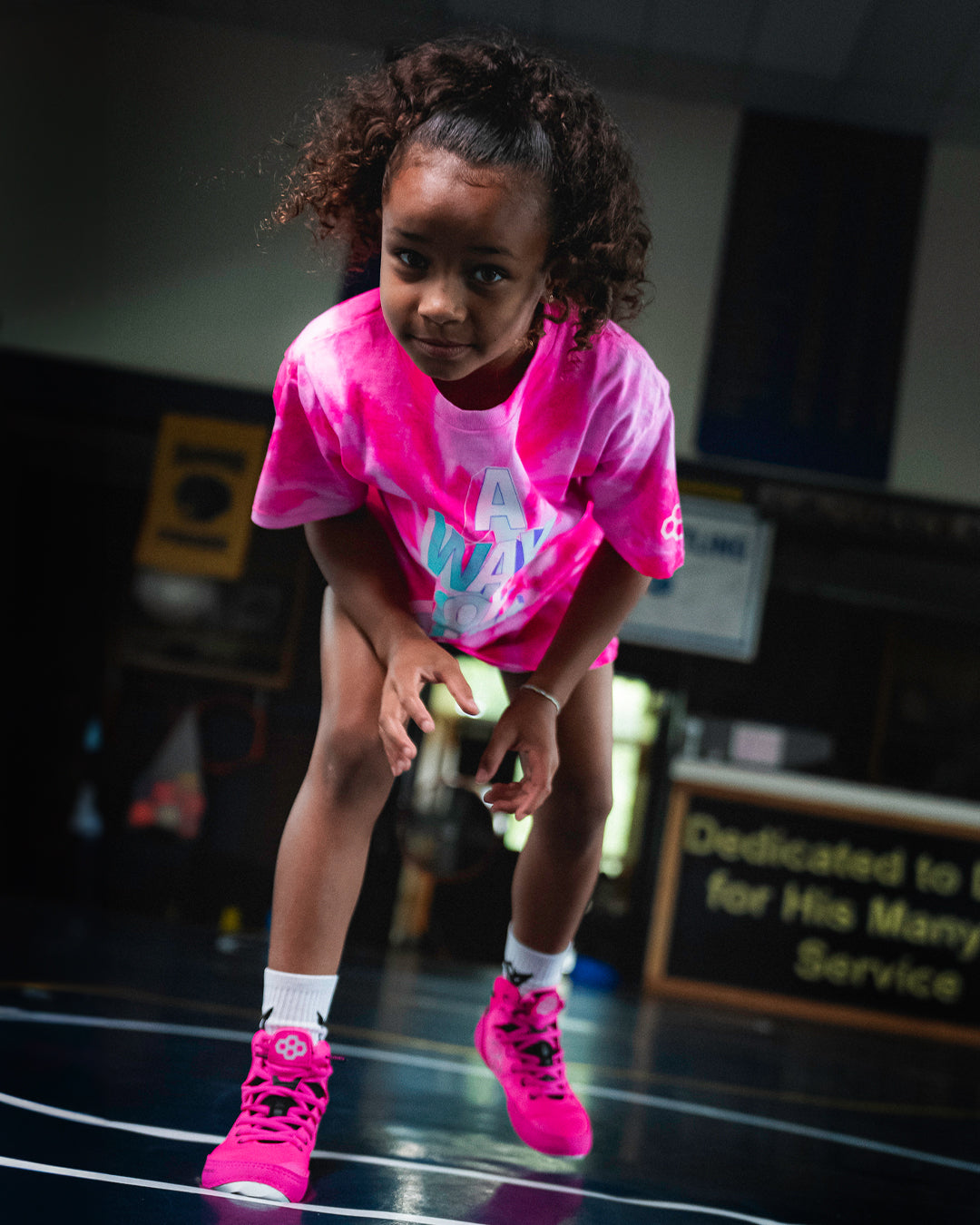 Youth wrestler Ora Burroughs in a pink RUDIS tie-dye shirt and bright pink RUDIS wrestling shoes getting into stance on the mat, showing focus, energy, and the future of girls’ wrestling.