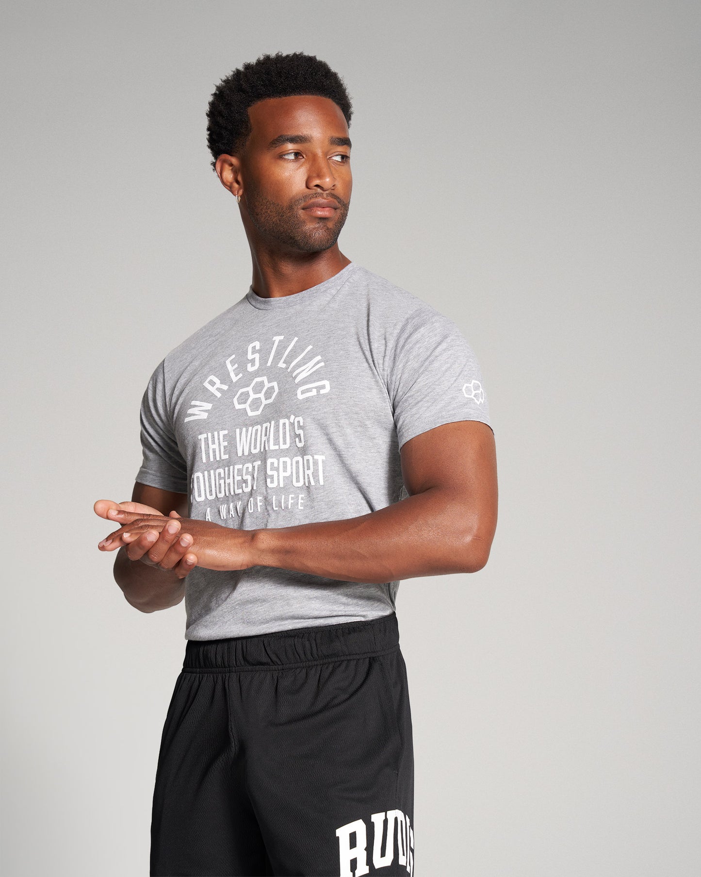 A muscular man wearing a gray wrestling t-shirt and black athletic shorts stands in a contemplative pose showcasing his confidence and readiness for action