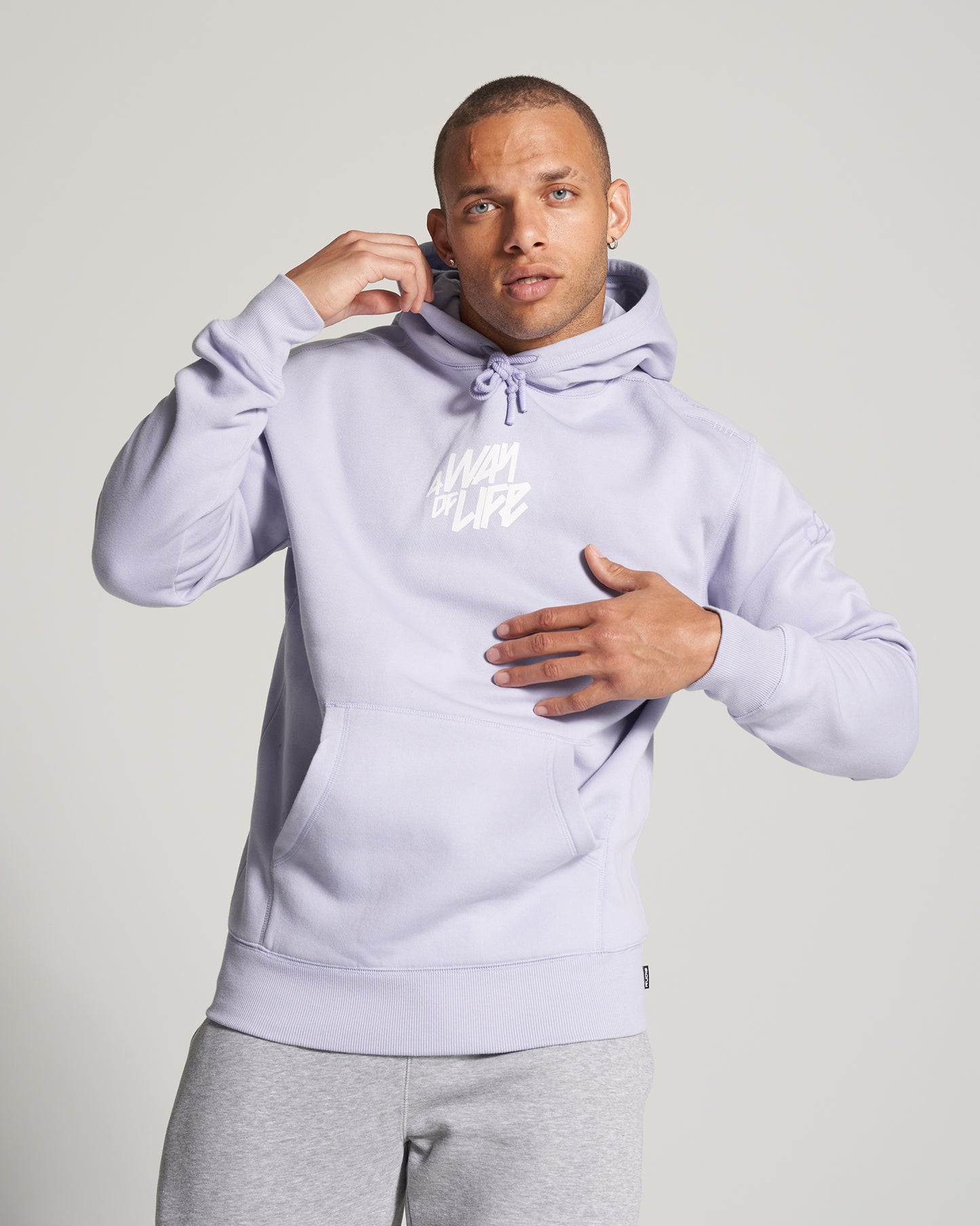 A young man poses confidently in a light purple hoodie with the phrase A WAY OF LIFE printed on the front paired with gray sweatpants showcasing a casual athletic style