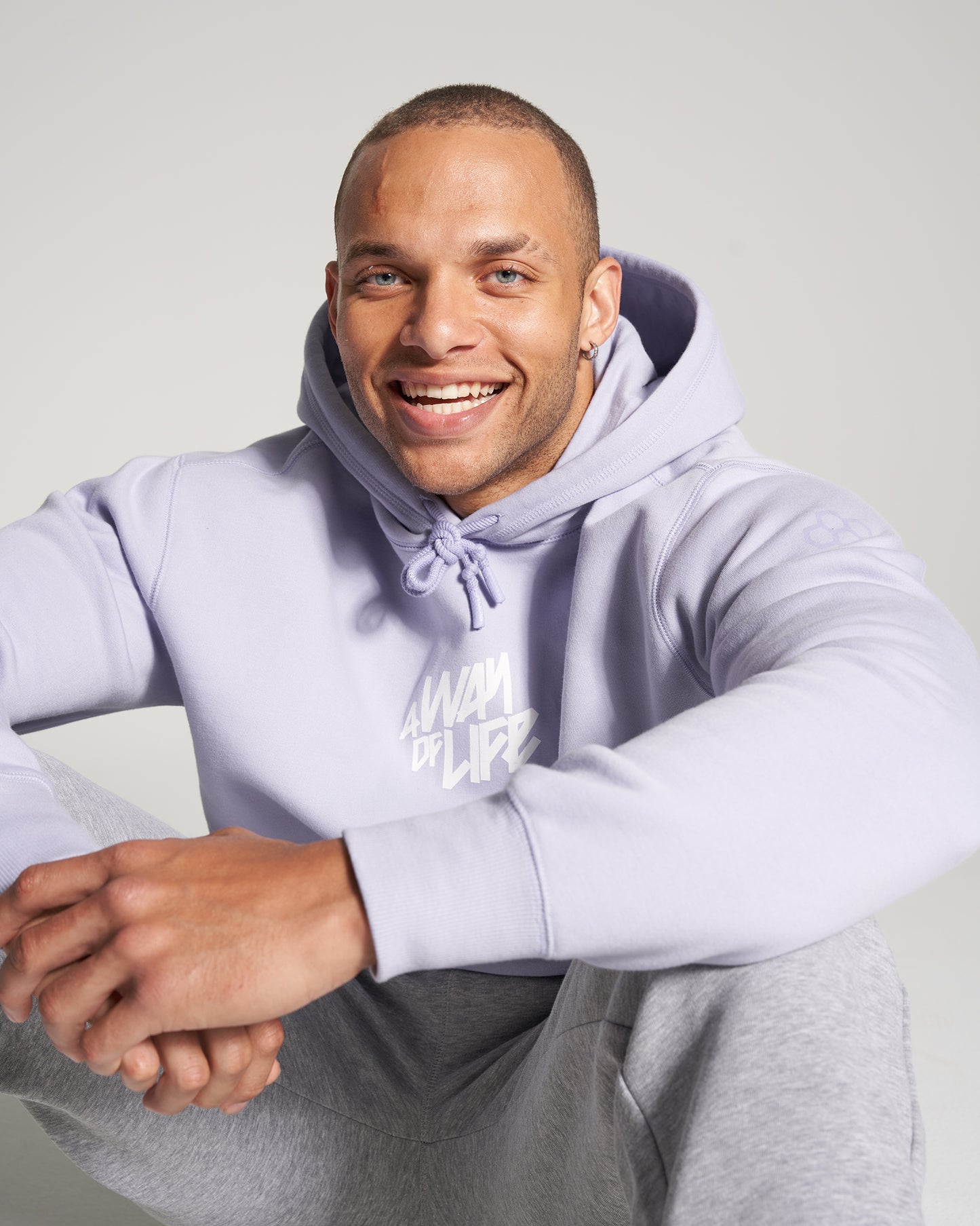 A smiling man sitting in a light purple hoodie and gray sweatpants exuding a relaxed and approachable demeanor