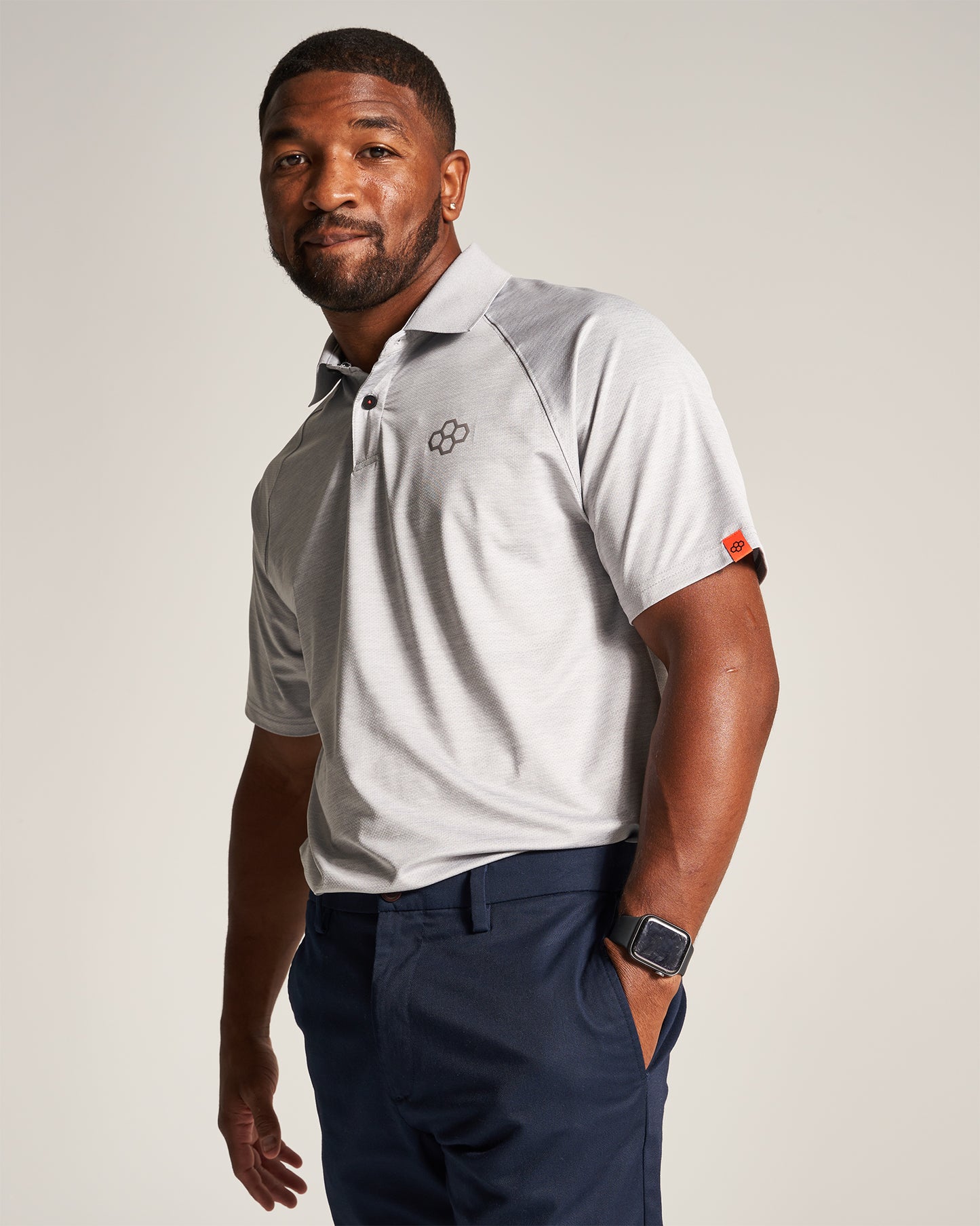 A man poses confidently in a light gray polo shirt paired with dark navy pants and a smartwatch against a soft beige background