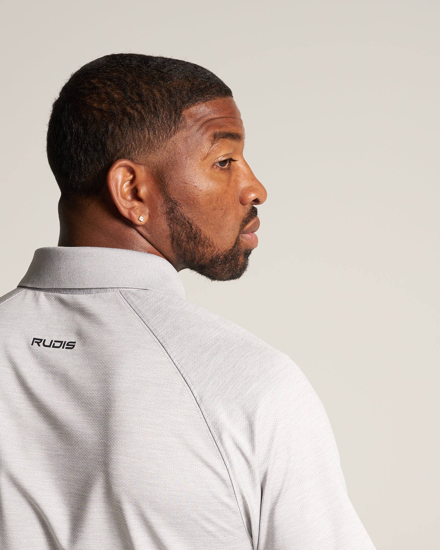 A man with a well-groomed beard wearing a light gray shirt with a collar and the brand name RUDIS visible on the back looking over his shoulder against a neutral background