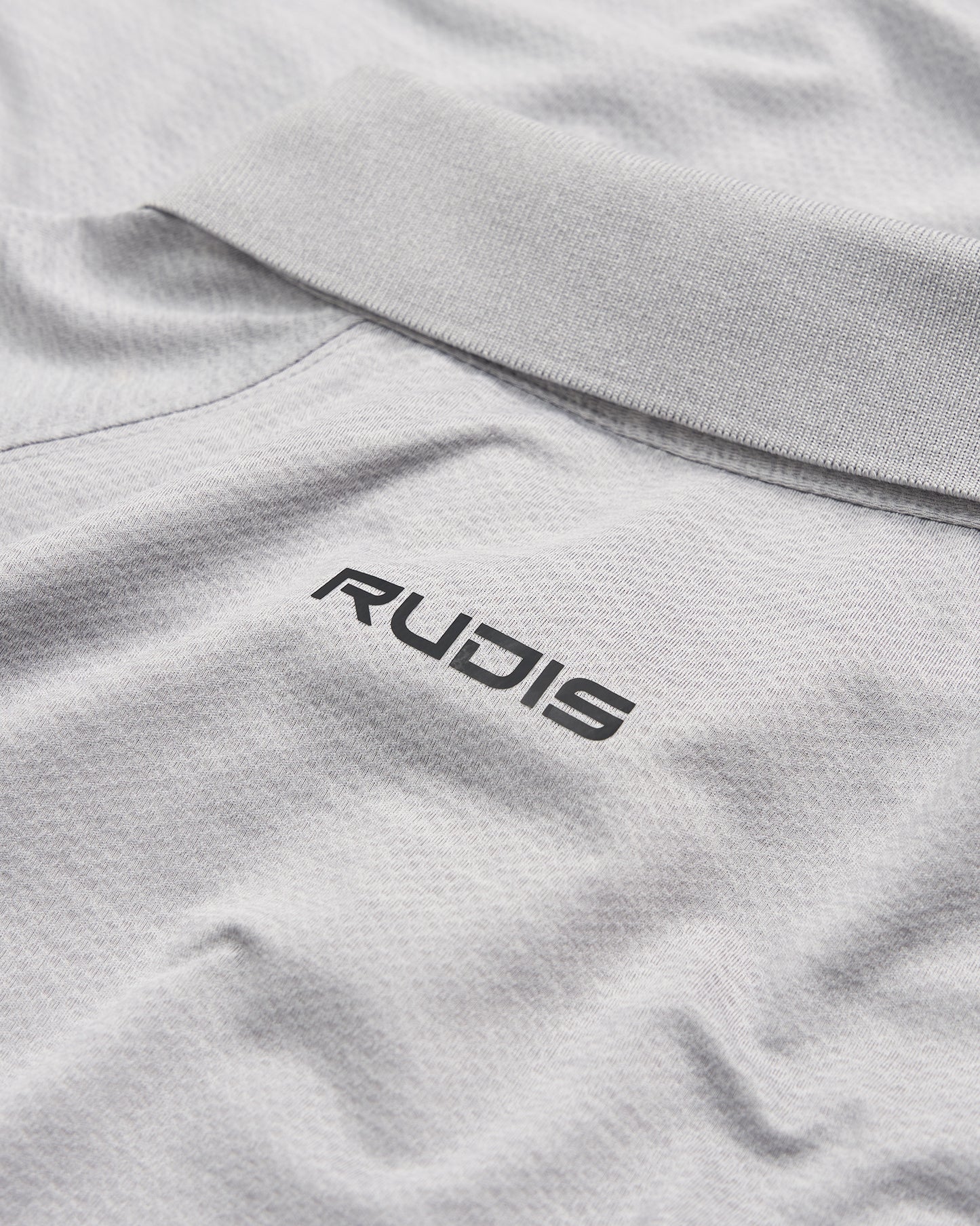 A close-up view of a light gray polo shirt featuring a black RUDIS logo on the collar area emphasizing a stylish and sporty aesthetic