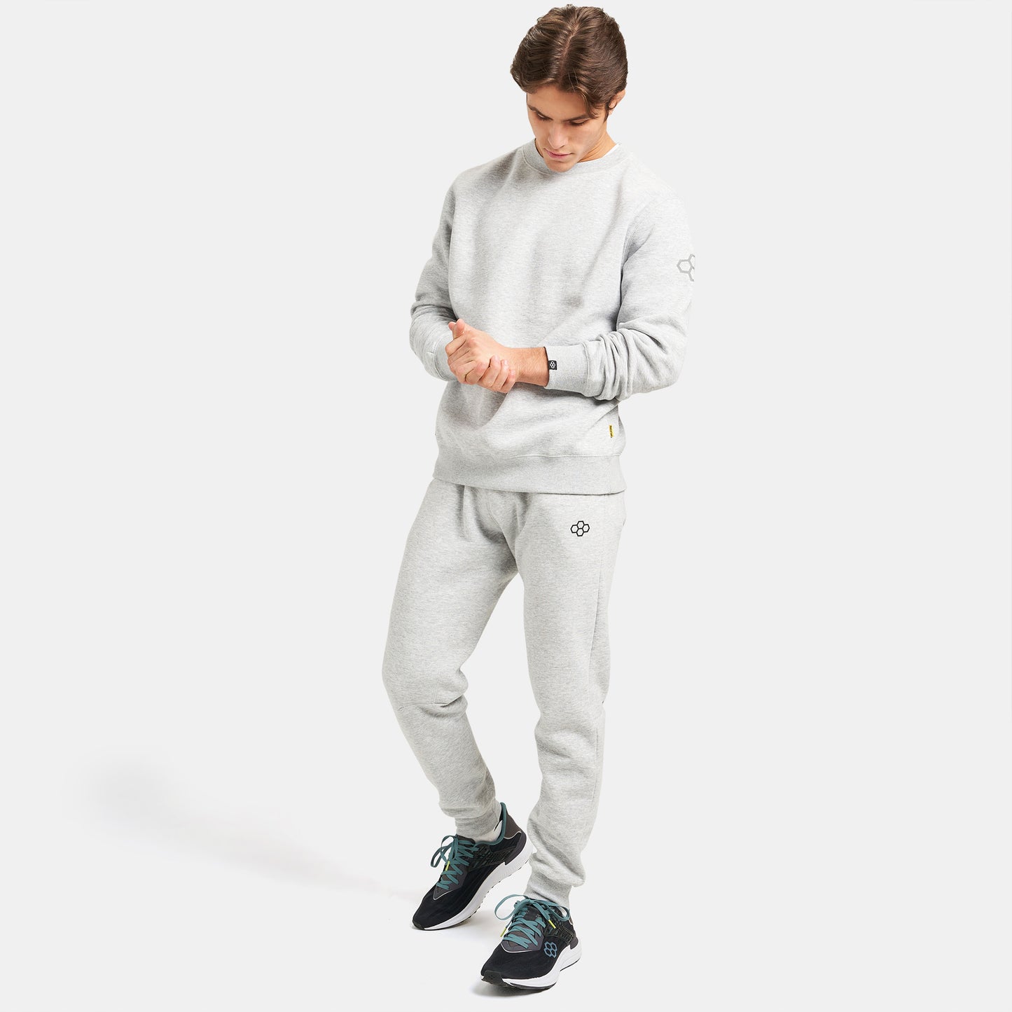 A young man dressed in a light gray athletic sweatshirt and matching sweatpants standing confidently with his hands on his wrists complemented by black sneakers featuring turquoise laces