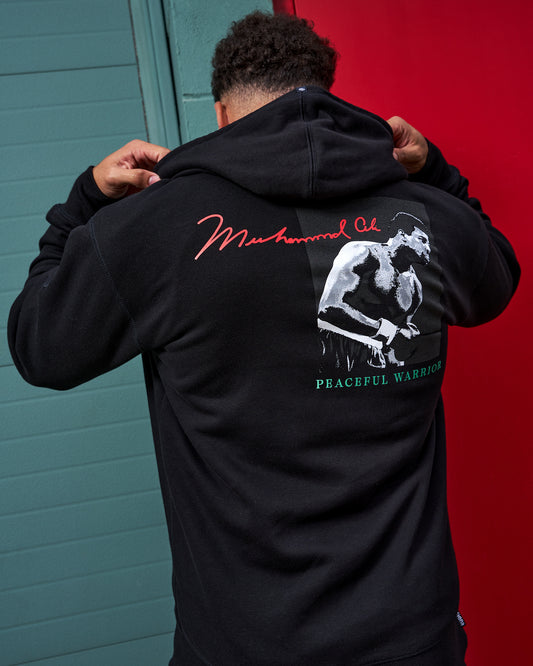 Person wearing a black hoodie with 'Muhammad Ali' design on the back against a red and blue wall.