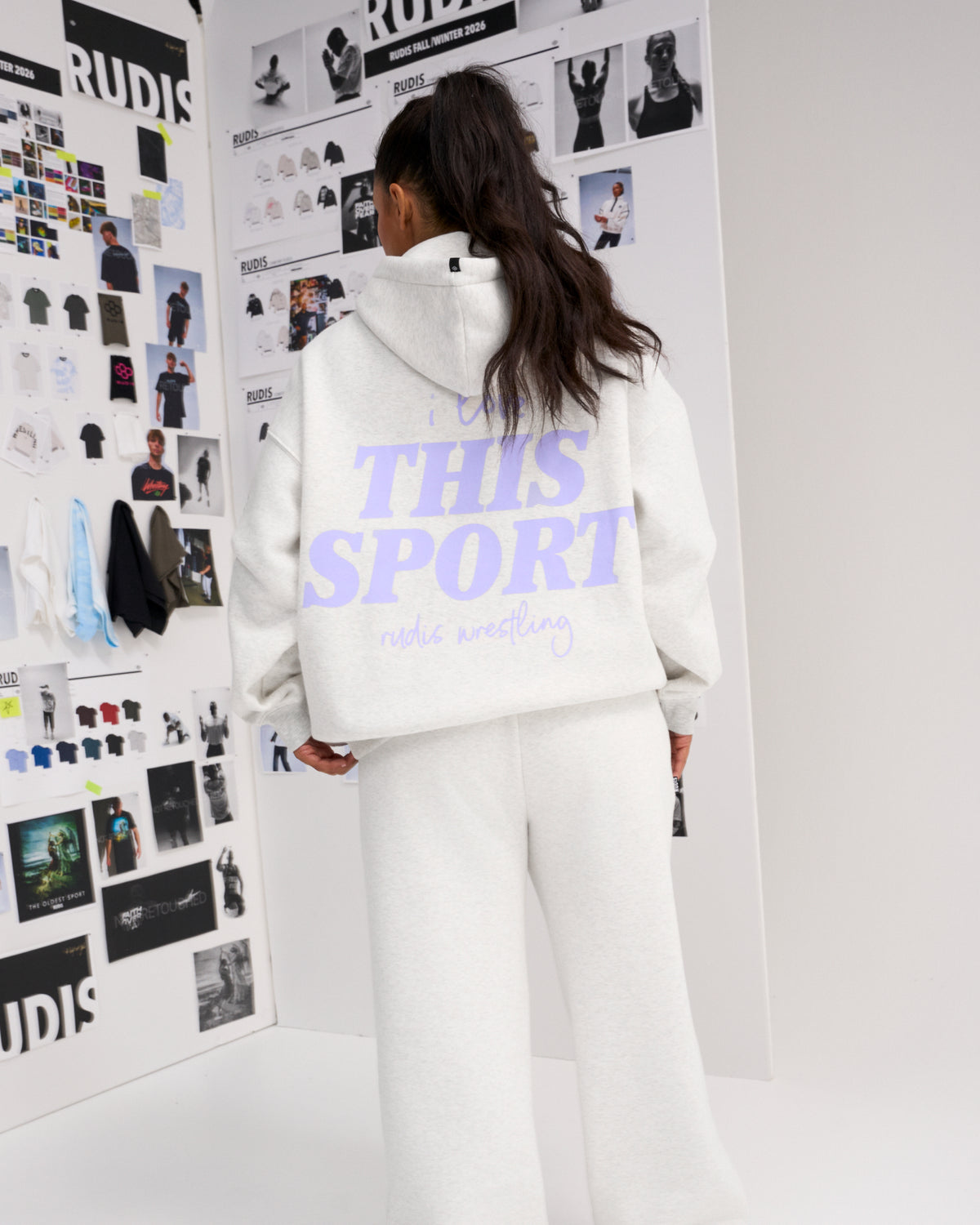 Rear view of a female model wearing the RUDIS Love This Sport Relaxed Comfort Fleece Hoodie in Heather Silver with lavender back graphics and matching sweatpants.