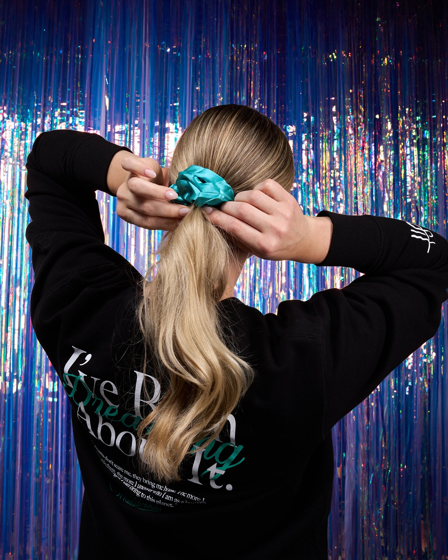 Person tying a teal scrunchie in their blonde hair against a colorful curtain background