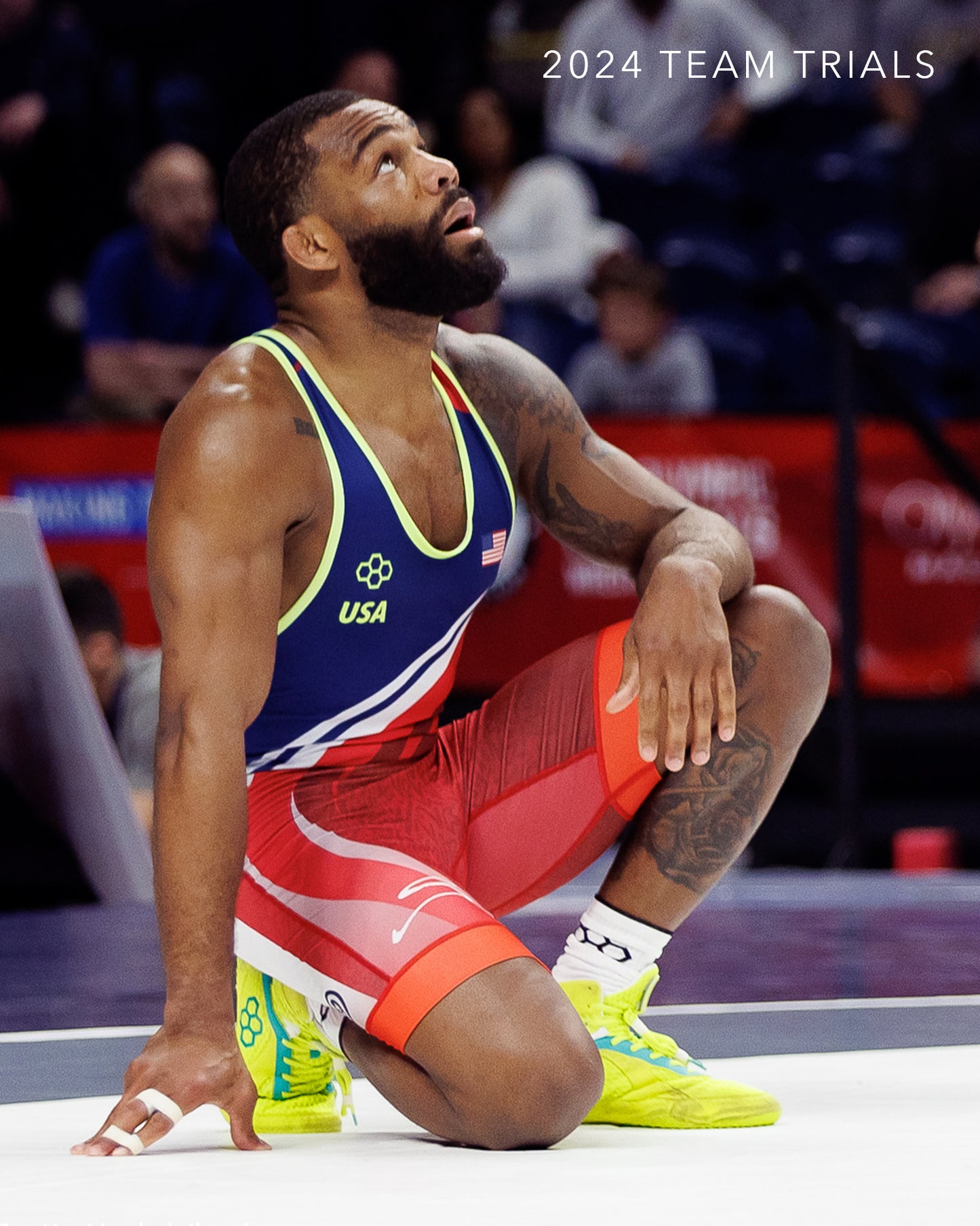 A male wrestler in a blue and red uniform kneels on a mat looking upwards with a focused expression during the 2024 Team Trials event
