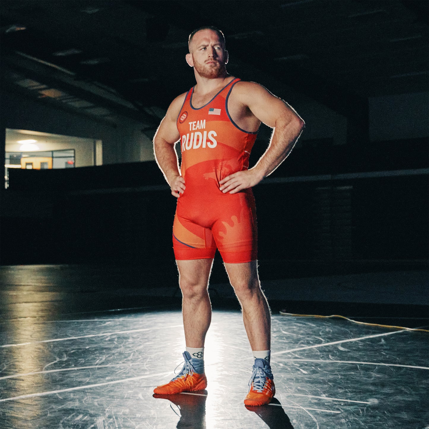 A muscular wrestler in a vibrant red uniform stands confidently on a wrestling mat showcasing his strong physique and determination