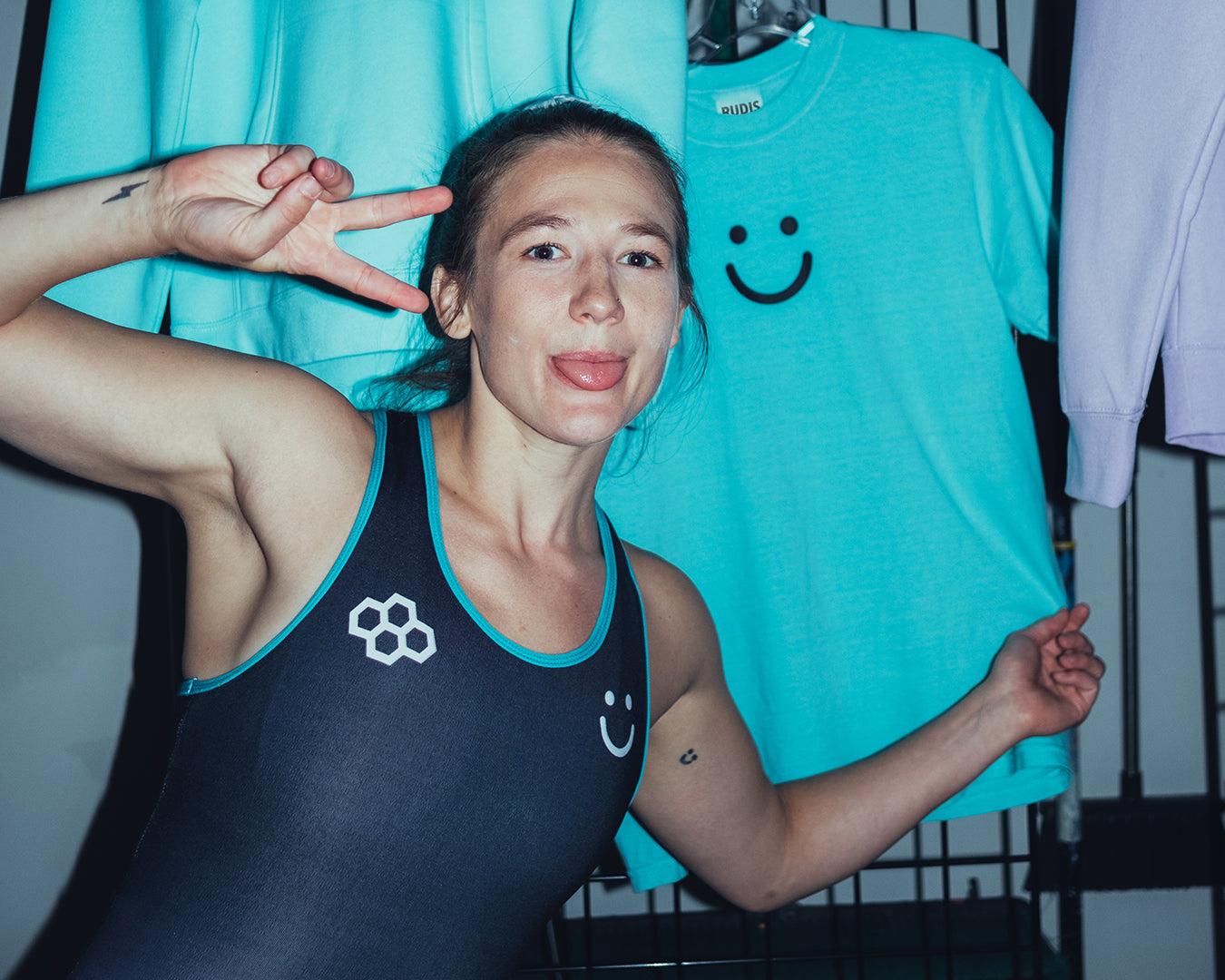 Team USA wrestler Sarah Hildebrandt showing off her signature RUDIS apparel line, posing playfully in a custom RUDIS singlet with smiley design in front of matching lifestyle clothing – blending personality, performance, and women’s wrestling style.