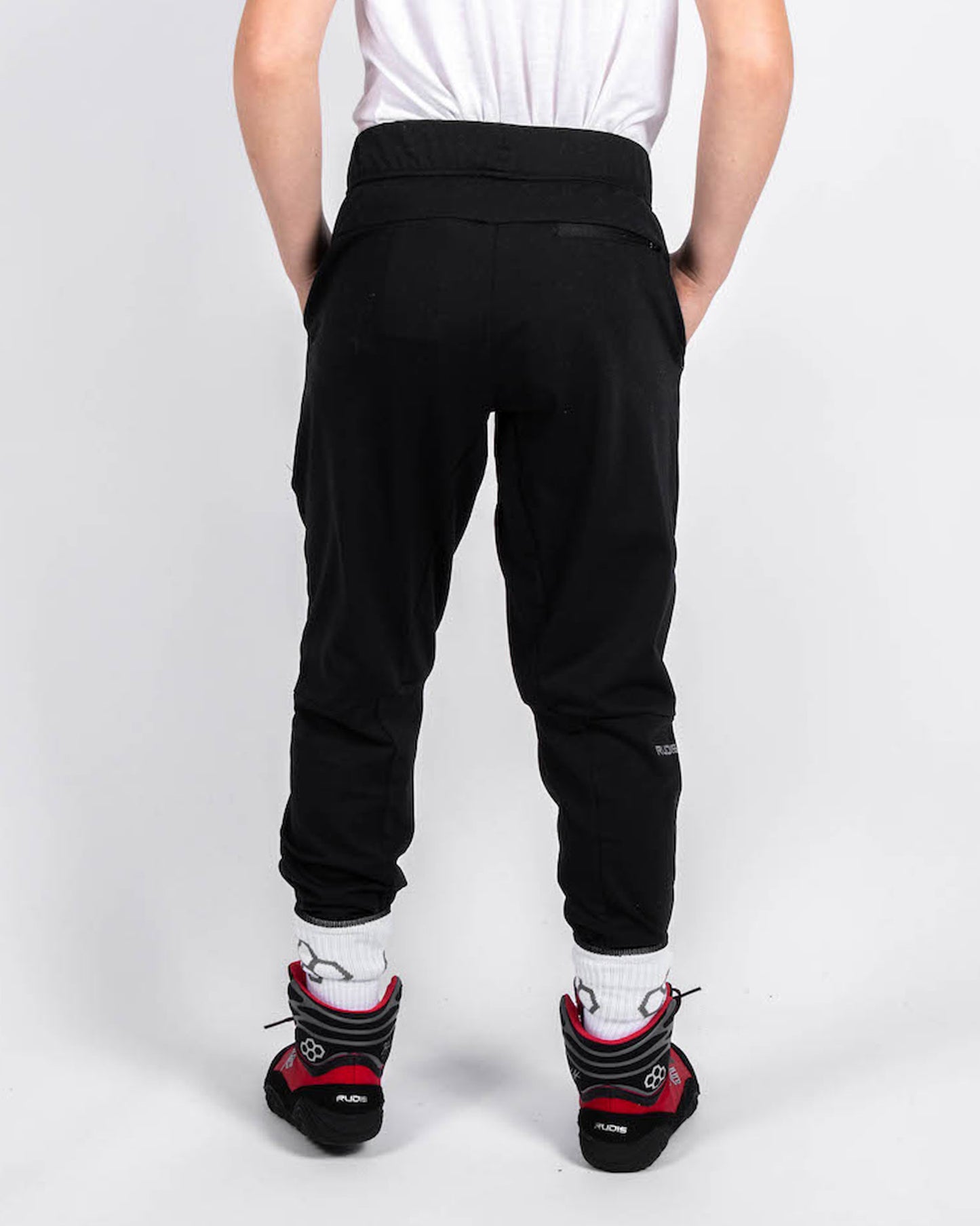 A young person wearing black sweatpants and red and black climbing shoes standing with their back facing the camera against a plain white background