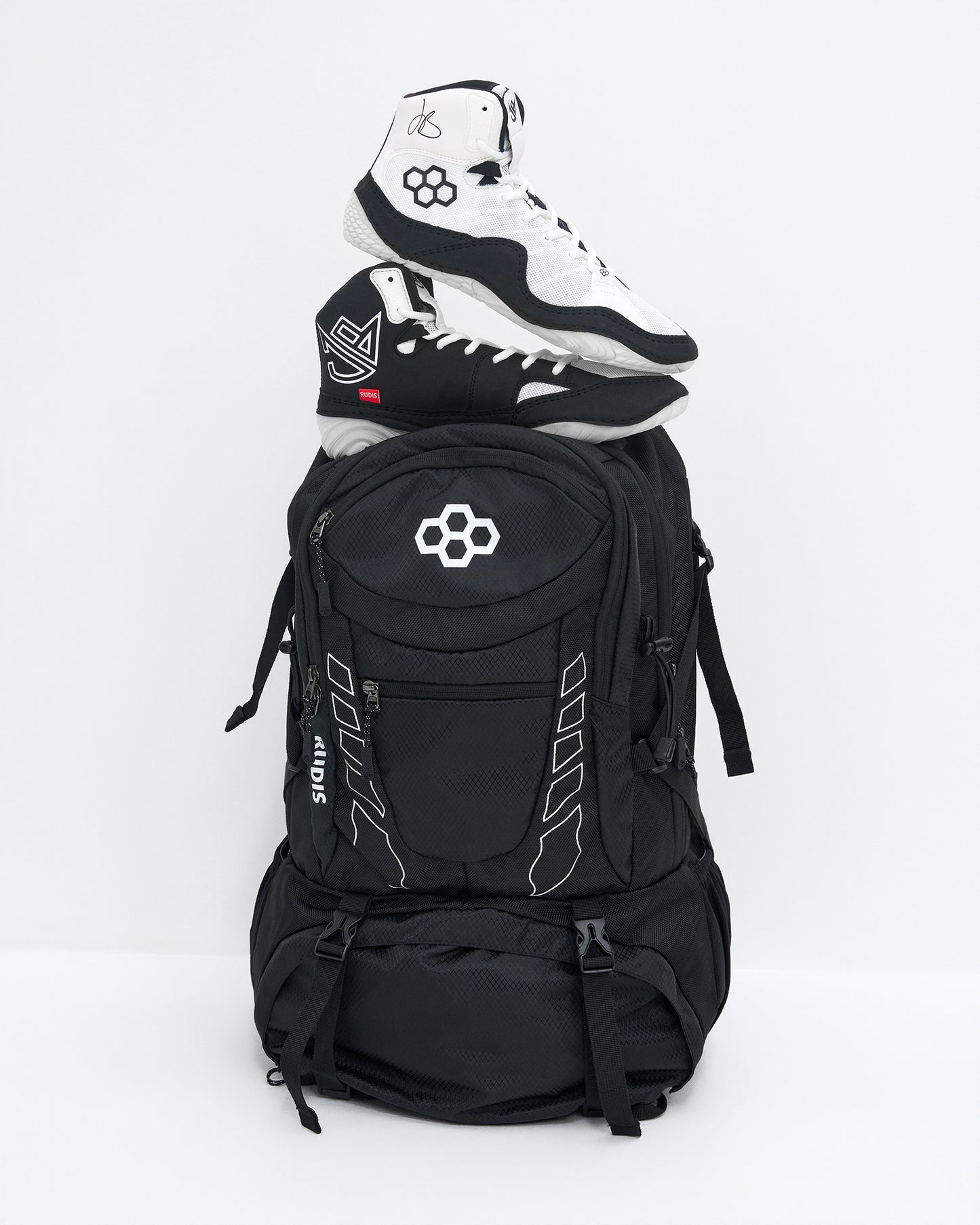 A black backpack with white graphic details and a white geometric logo on the front pocket, styled with a pair of black and white high-top wrestling shoes stacked on top, set against a plain white studio background.