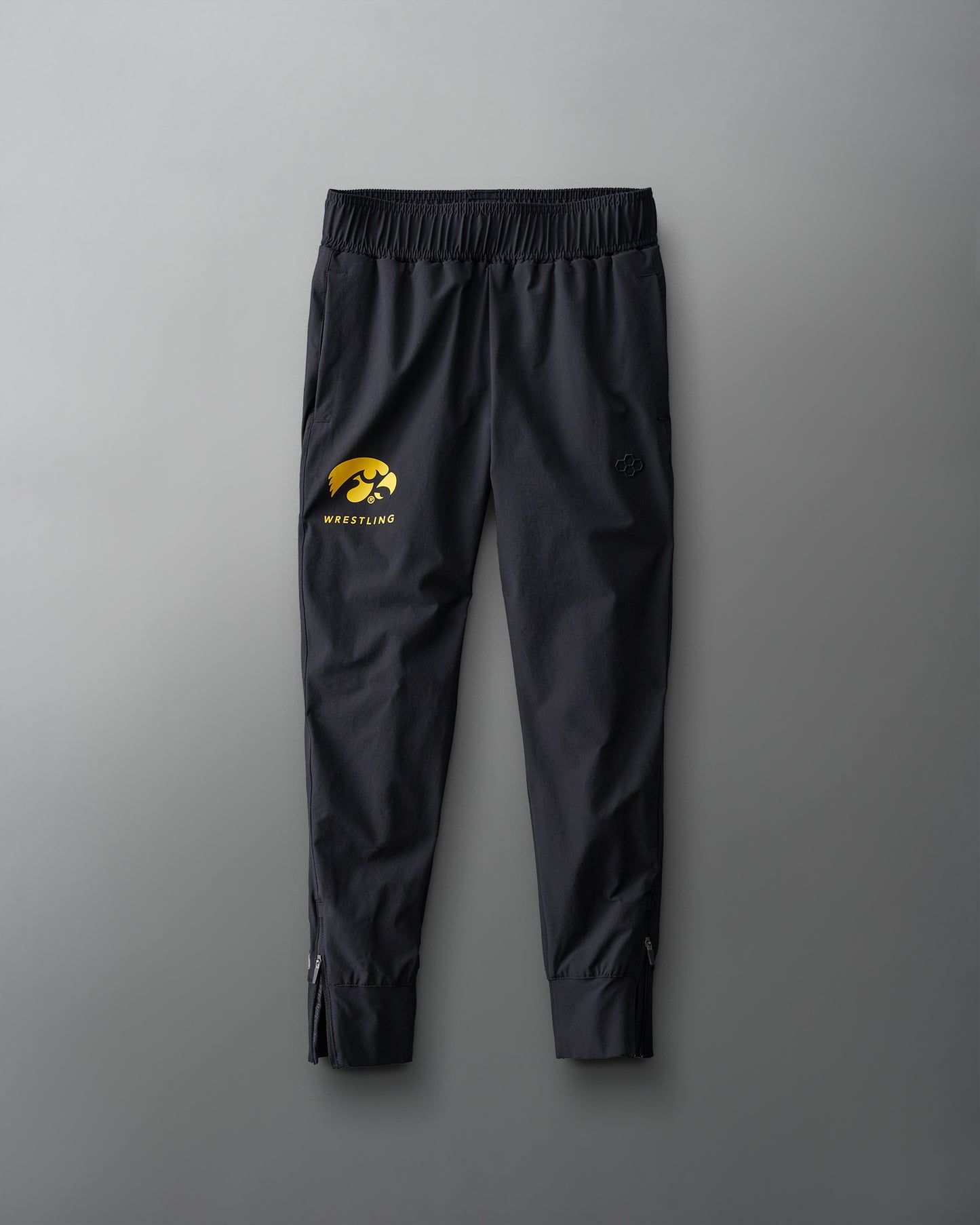 Youth athletic pants in black with a gold iowa hawkeyes wrestling logo on the left leg, laid flat on a grey background.