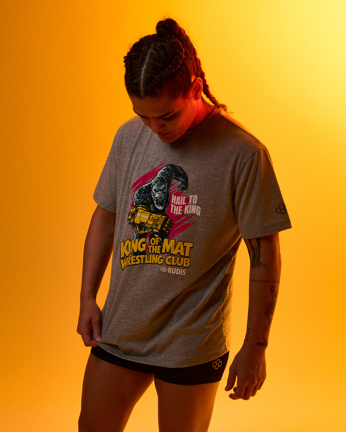 A model wearing a heather gray graphic t-shirt with a 'King of the Mat' wrestling design, posed against a warm orange-yellow gradient background.