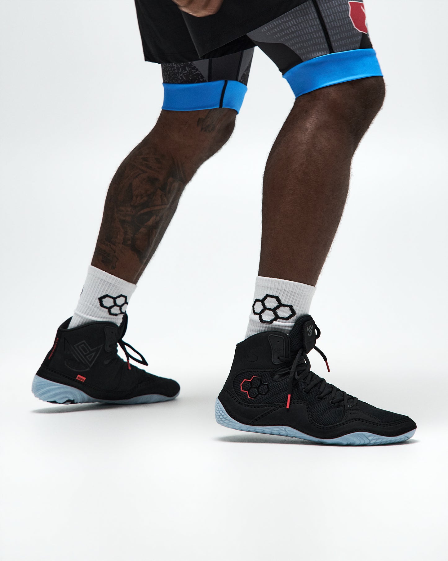 Black high-top athletic Wrestling shoes with light blue soles, worn with white crew socks and black athletic shorts with blue compression bands, shown on a model's legs against a white studio background.