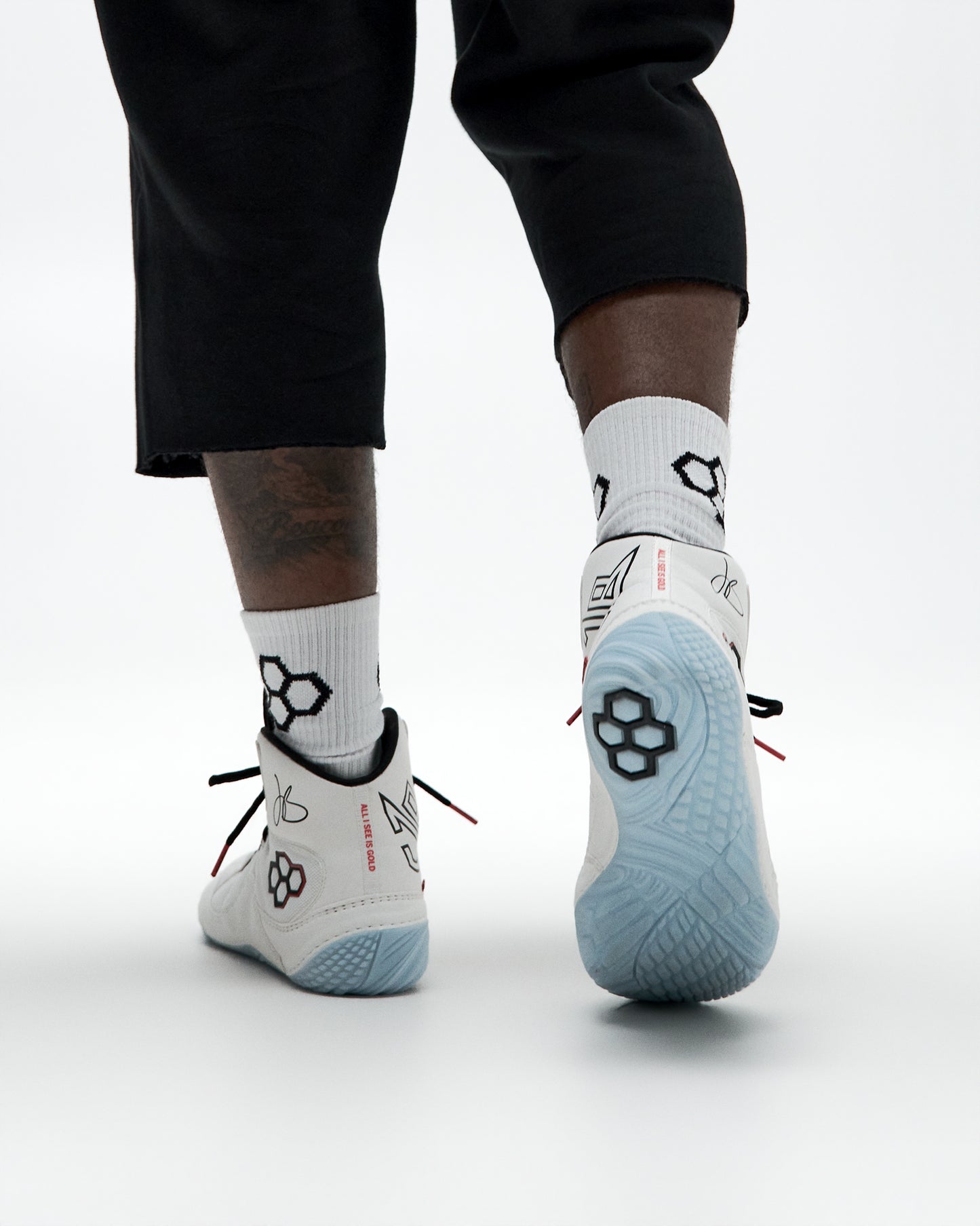 White and light blue Wrestling shoes with black graphic patterns, worn with white crew socks and black pants, shown from a rear three-quarter angle on a light gray studio background.