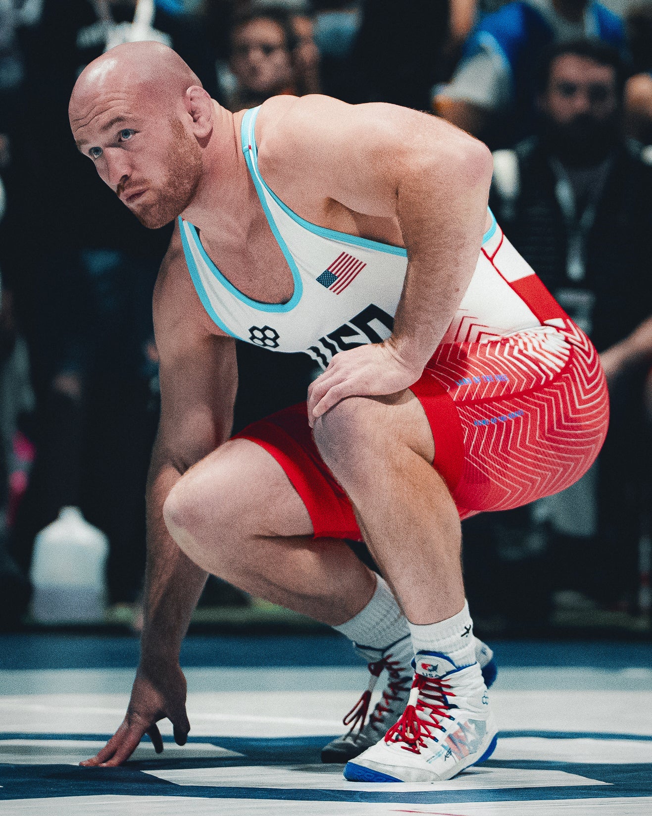 Kyle Snyder Wrestler wearing a USA singlet and RUDIS KS Infinity Edge Wrestling Shoes crouches on the mat in competition stance, with red and white wrestling shorts and red laces on white and blue shoes.