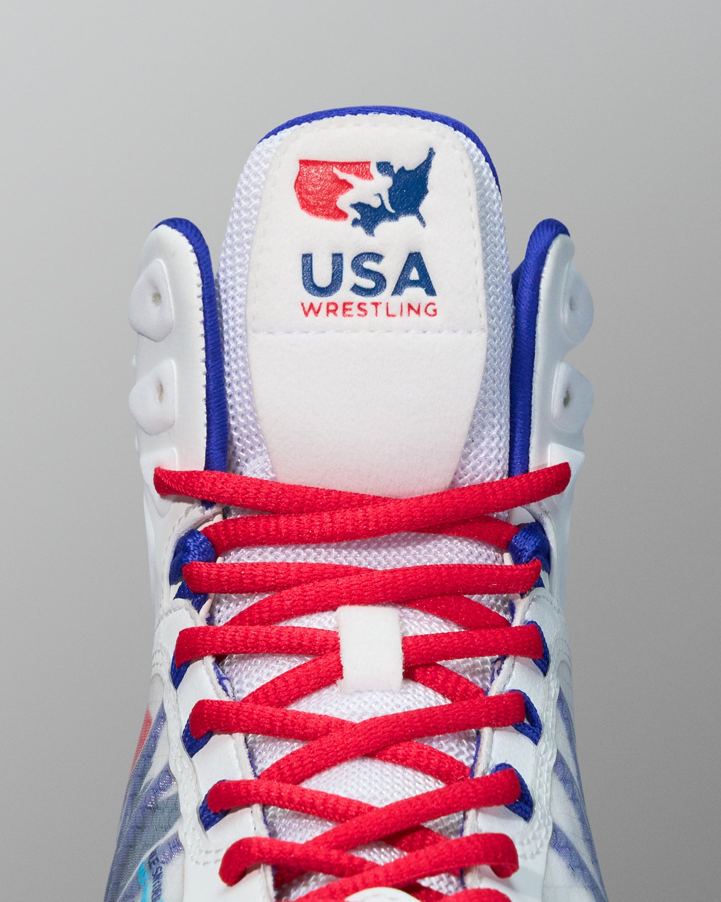 Close-up of the tongue and laces of a white RUDIS wrestling shoe featuring the USA Wrestling logo in red and blue stitching above red laces with blue accents.