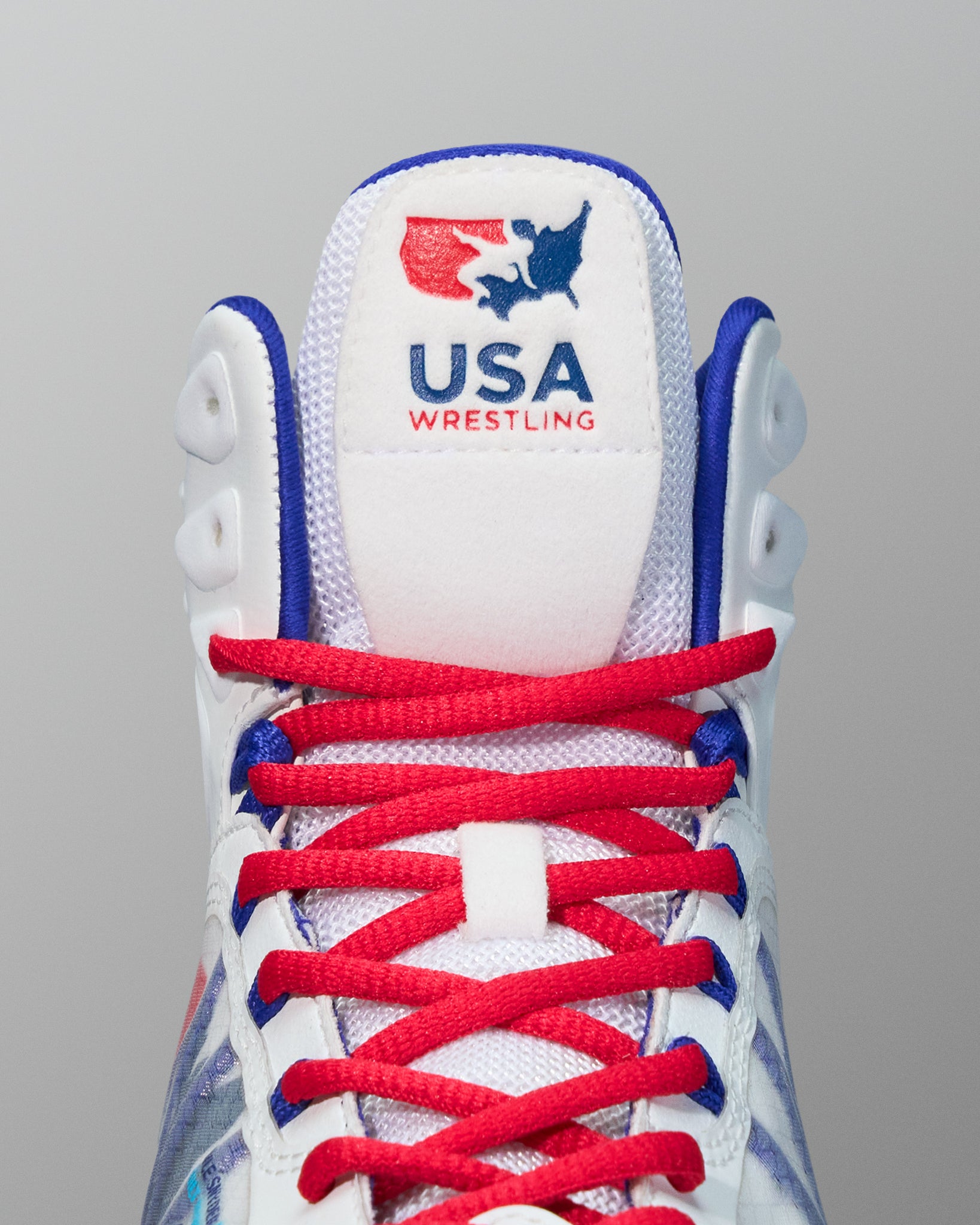 Close-up of the tongue and laces of a white RUDIS wrestling shoe featuring the USA Wrestling logo in red and blue stitching above red laces with blue accents.