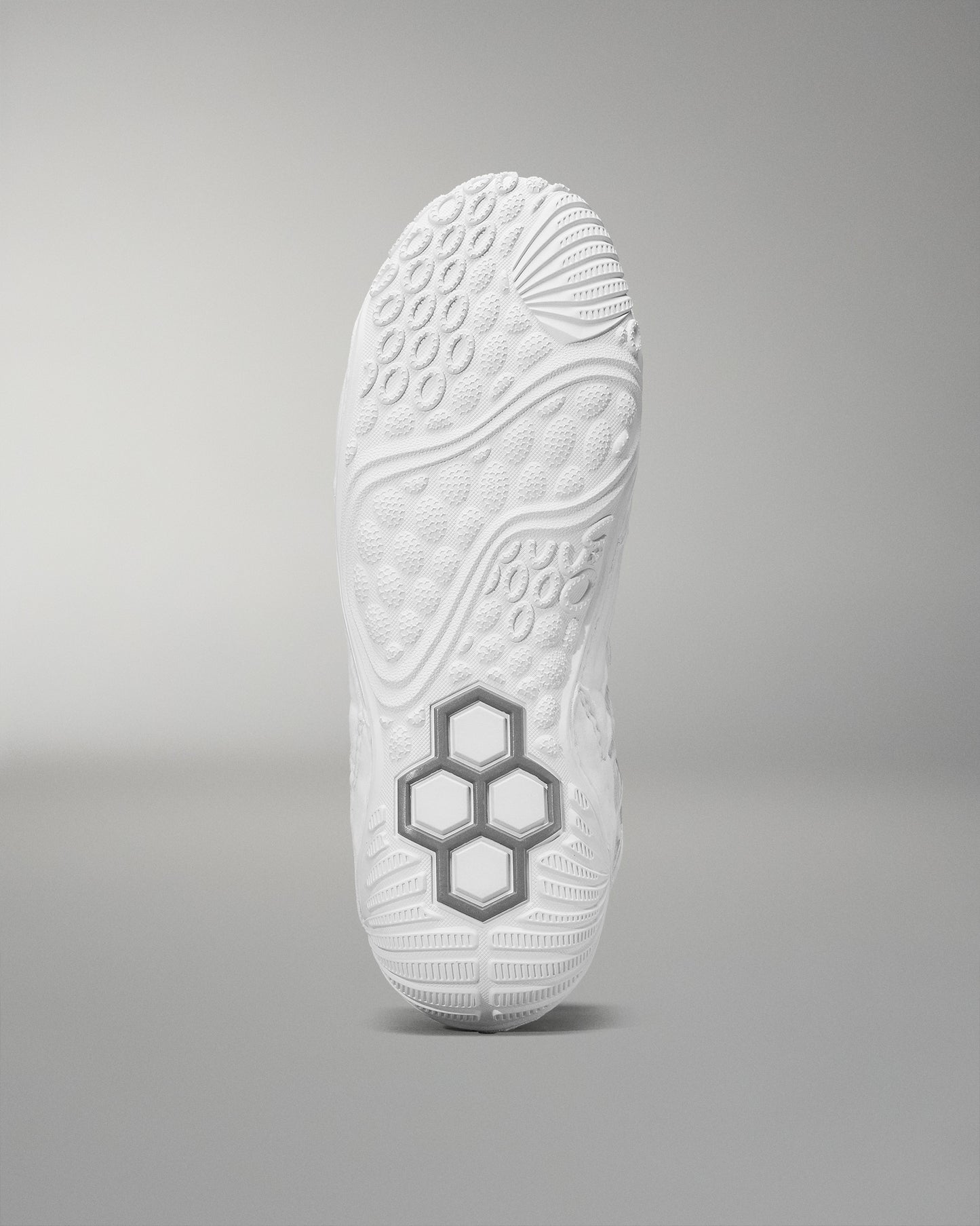 The sole of a white wrestling shoe, set against a soft gray background, showcases a textured design with raised patterns and a prominent gray hexagonal structure for improved grip.