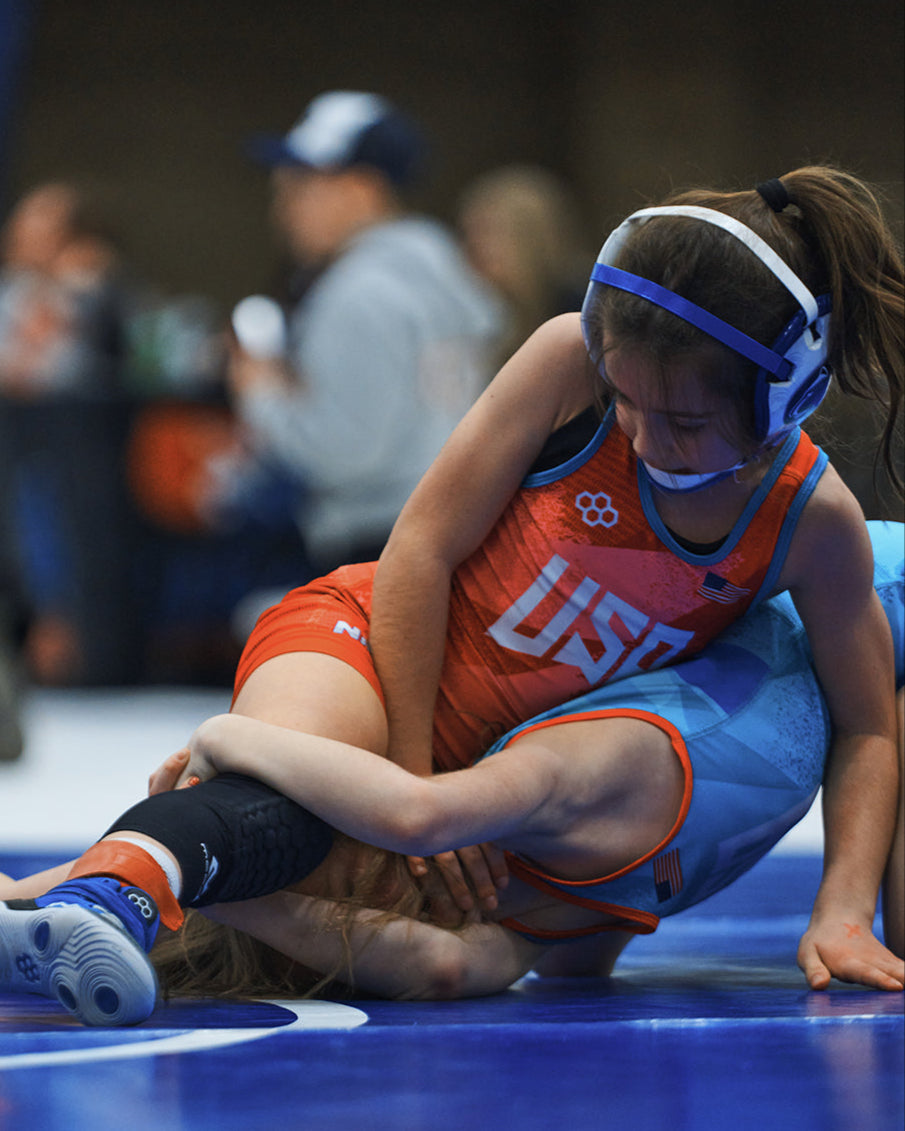 Young girl wrestler in a red USA Wrestling singlet competing at the Kids Folkstyle National Championship, controlling her opponent on the mat with focus and determination.