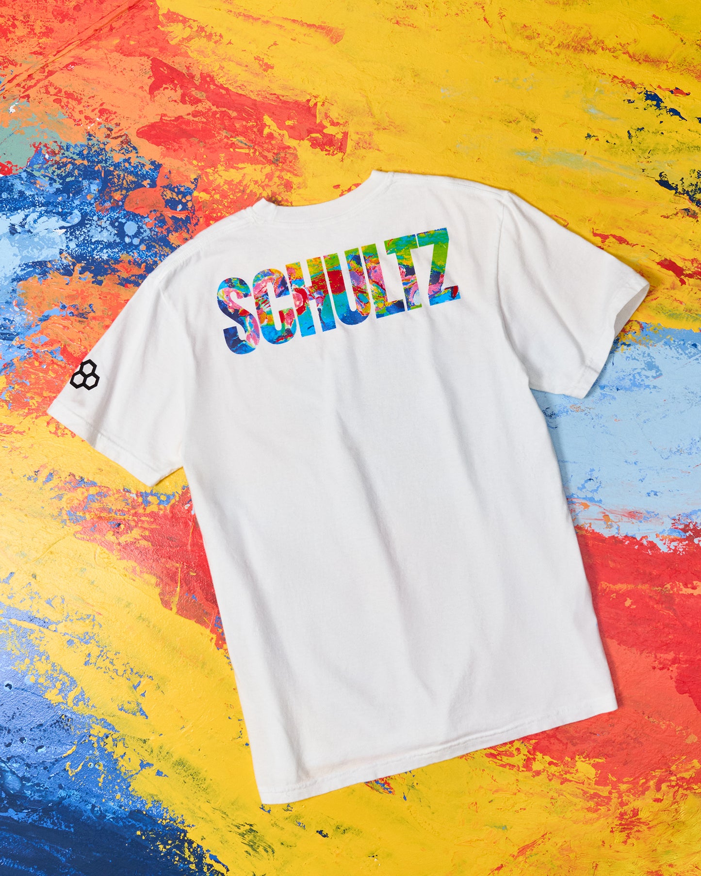 White t-shirt with colorful 'SCHULTZ' logo on a multicolored abstract background
