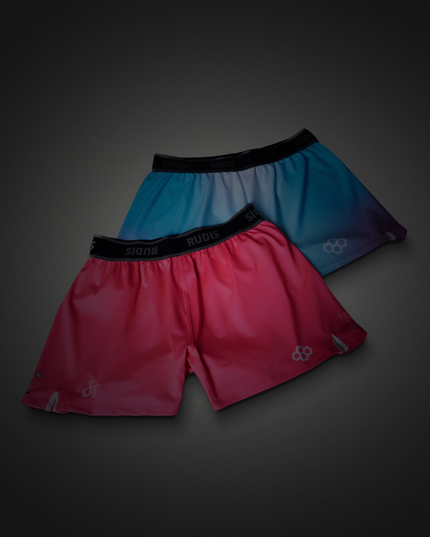 Two pairs of athletic shorts from the brand RUDIS, displayed against a dark gray gradient background. The front pair is a vibrant pink-to-red gradient with a black waistband, and the back pair is a blue-to-purple gradient with a black waistband.