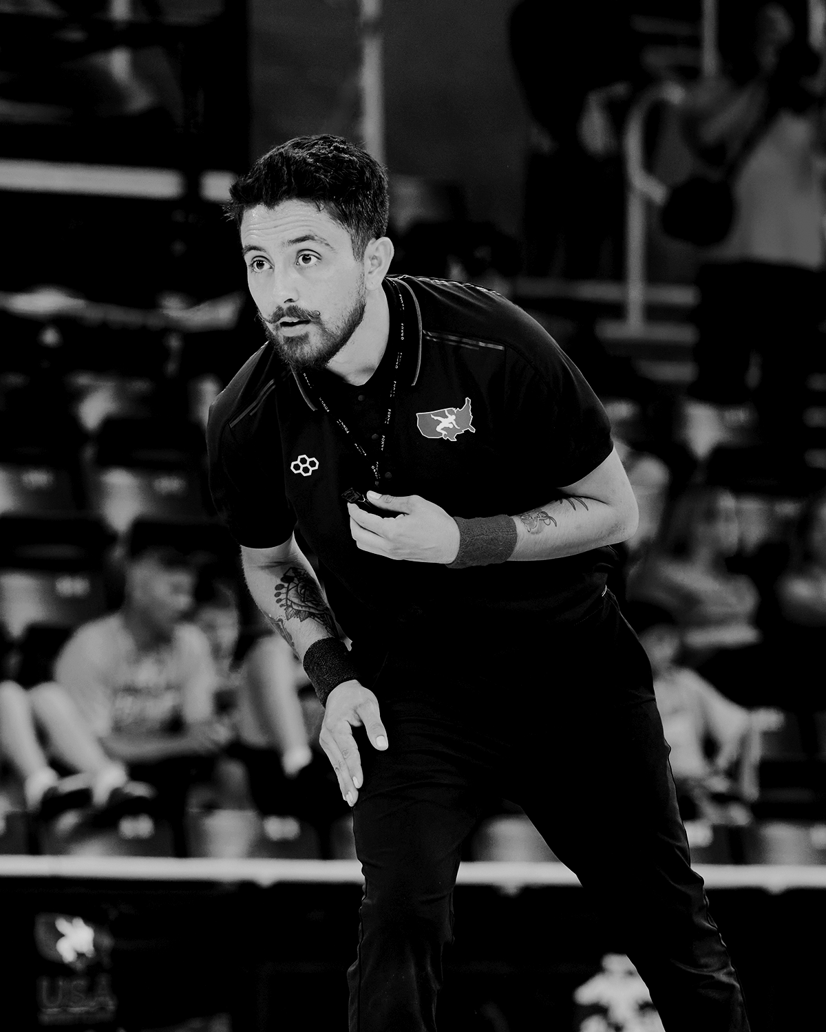 Black-and-white photo of a USA Wrestling referee in a RUDIS polo, leaning forward with intensity and focused mid-match, ready to make a call on the mat.