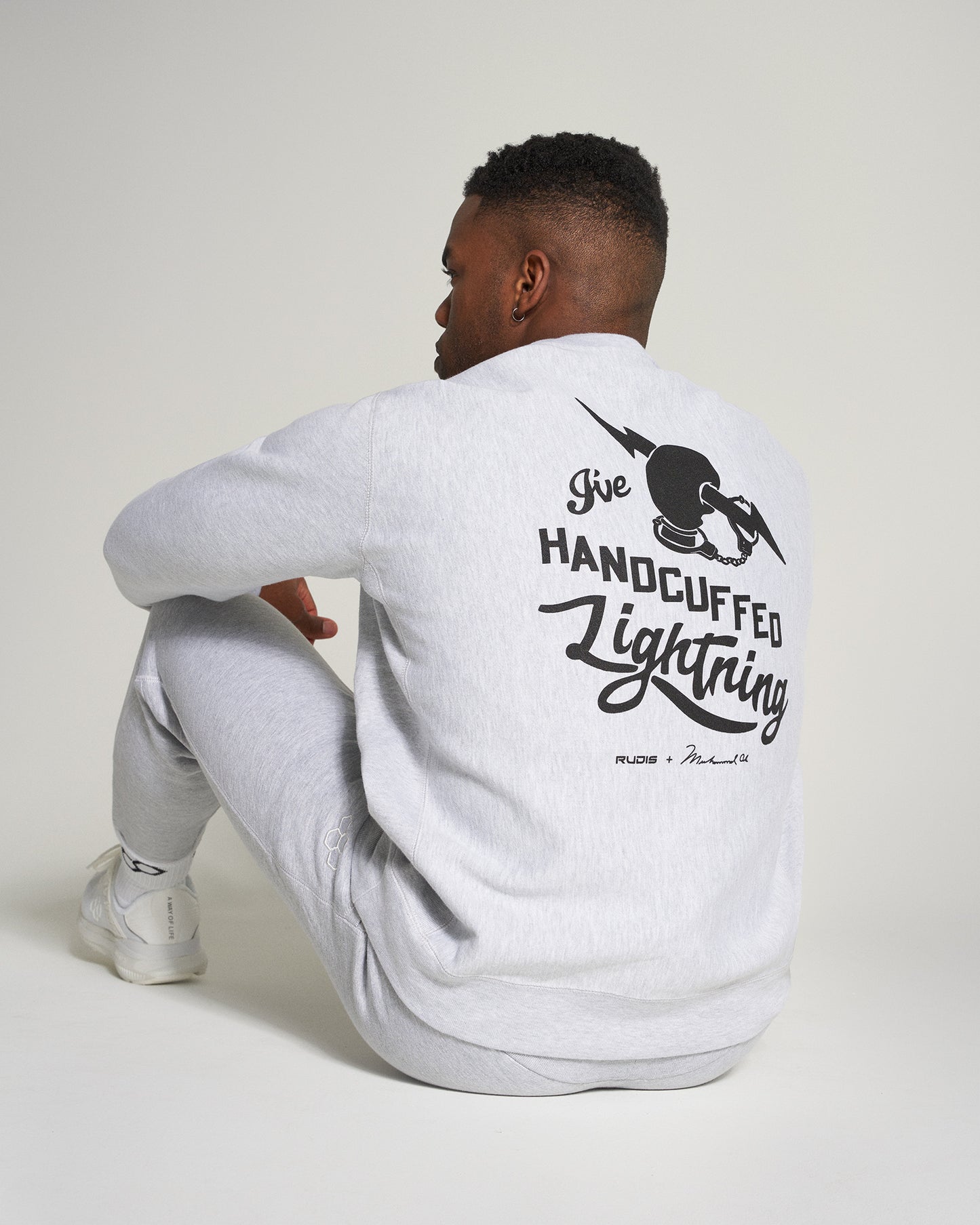 A model wearing a light gray sweatshirt with the phrase Ive Handcuffed Lightning printed on the back paired with matching gray sweatpants showcasing a casual streetwear style