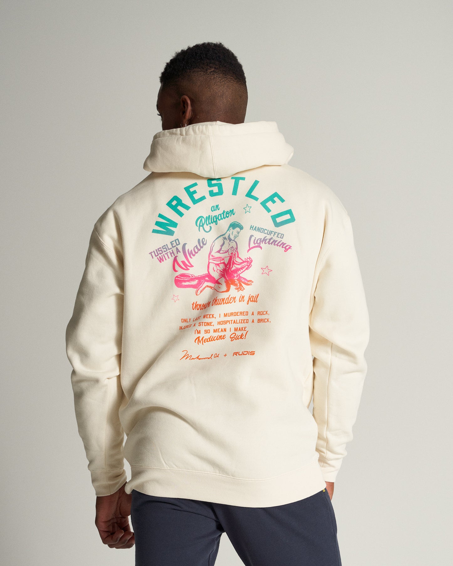 A person wearing a cream-colored hoodie featuring vibrant graphics including phrases about wrestling and humorously exaggerated feats