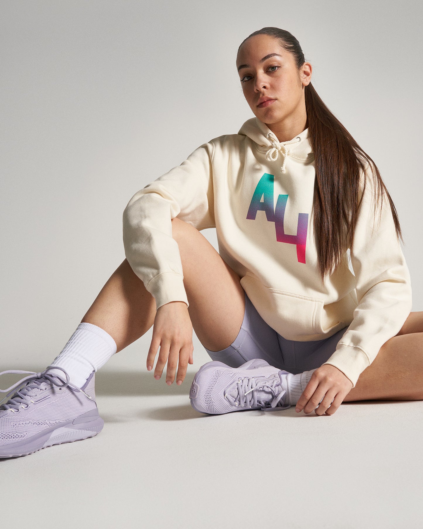 A young woman sits in a relaxed pose wearing a light beige hoodie featuring a colorful ALY logo paired with light gray shorts and lavender athletic shoes