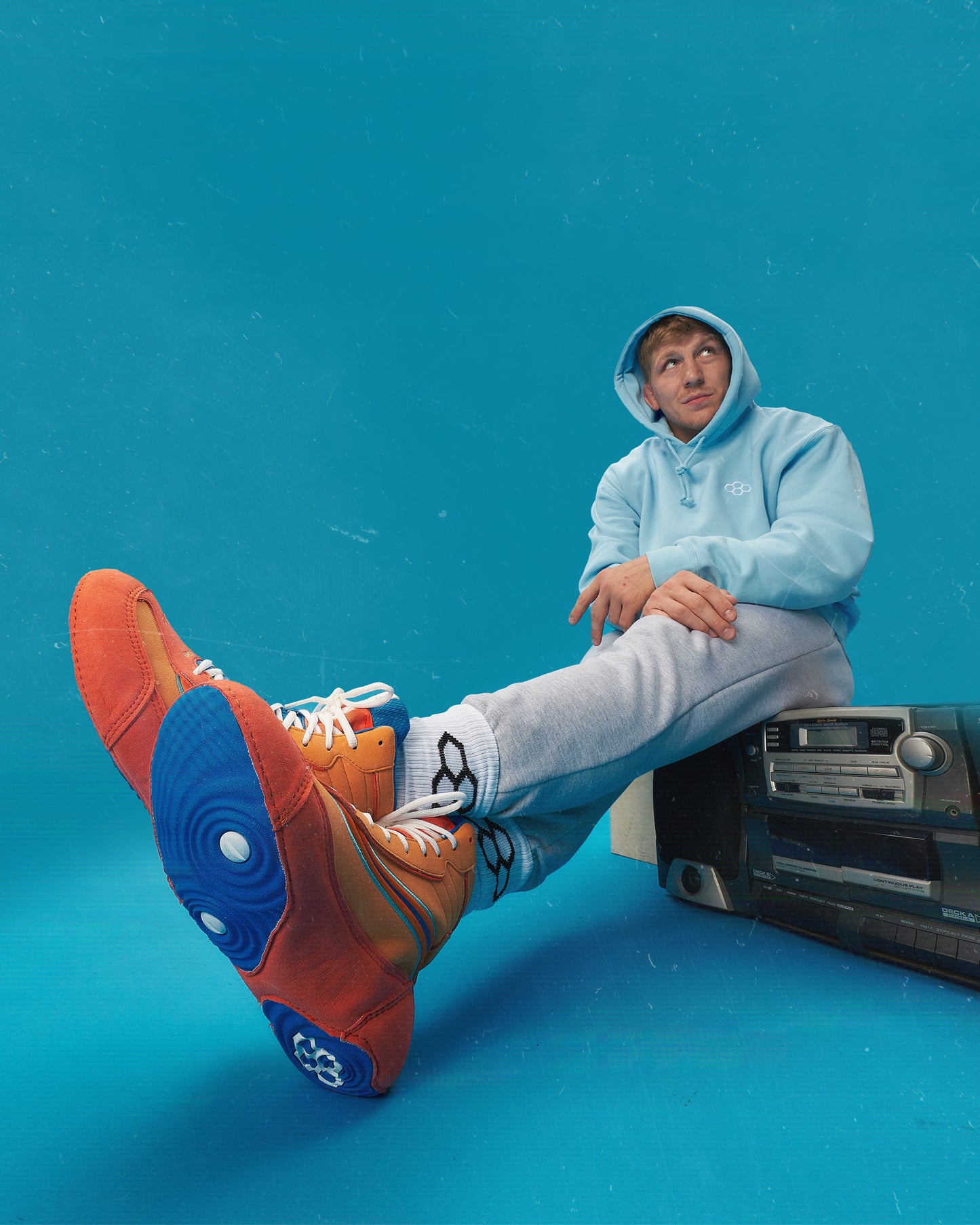A person wearing a light blue hoodie and gray sweatpants sits next to a vintage cassette player showcasing vibrant orange and blue shoes
