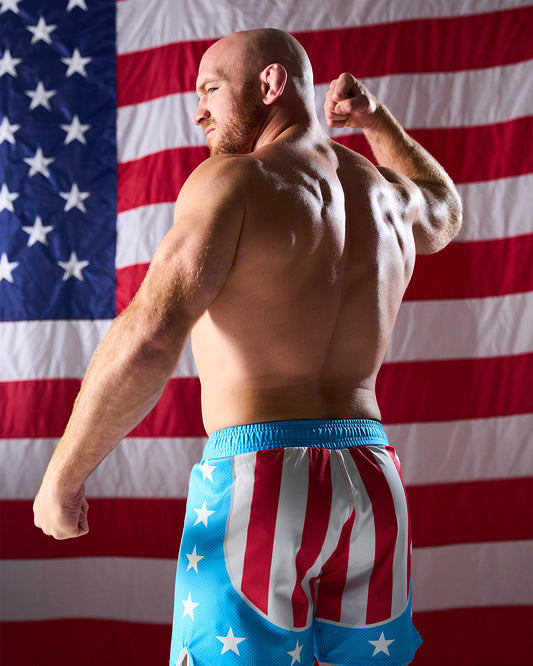 Man flexing muscles wearing American flag-themed shorts with a U.S. flag in the background