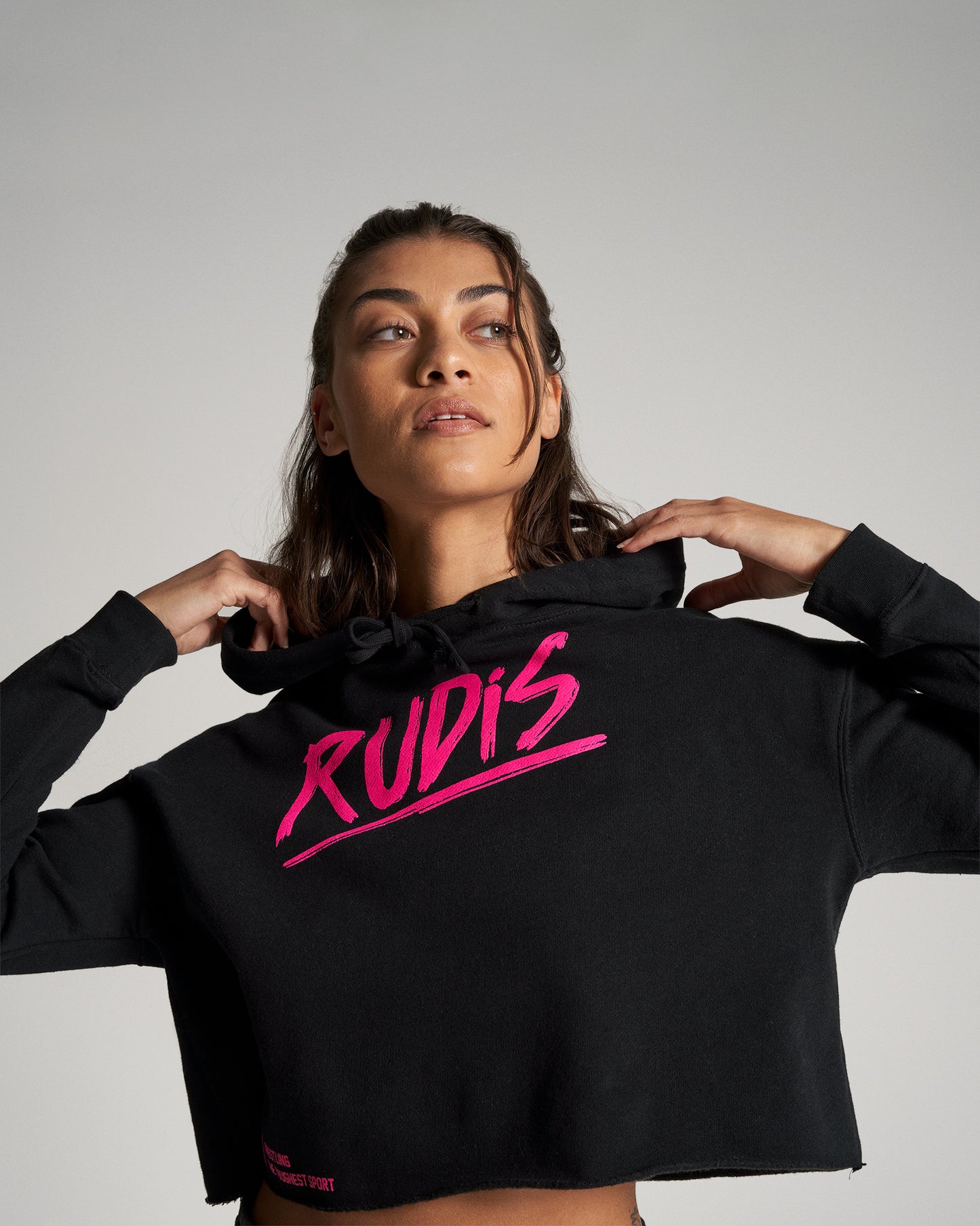A close-up portrait of a woman in a stylish black cropped hoodie featuring bright pink lettering that reads RUDIS set against a neutral background