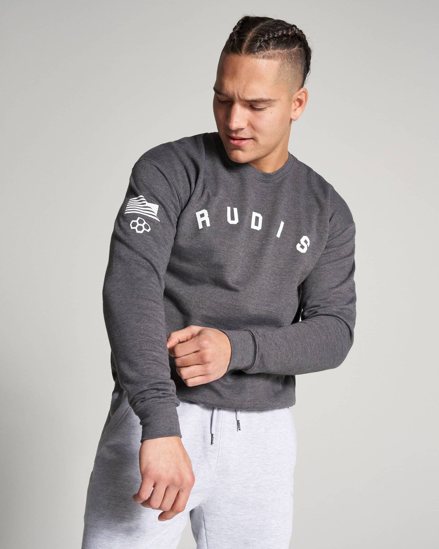 A male model in a gray sweatshirt with RUDIS printed on the front wearing light gray pants and adjusting the sleeve of his shirt