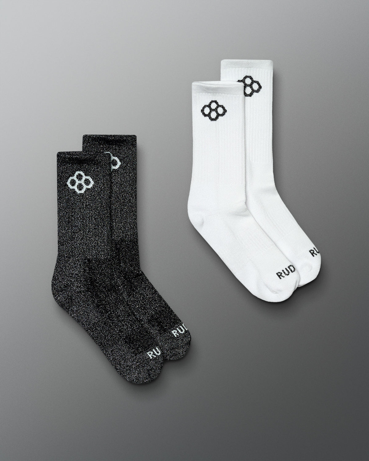 Two pairs of crew socks on a smooth gray background: one pair is black with a white logo and the other pair is white with a black logo. The word 'RUD' is visible near the toes.