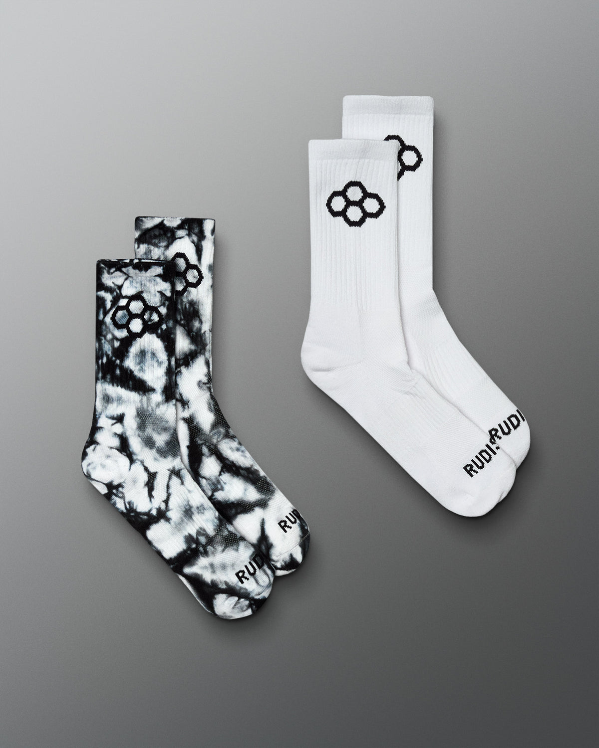 Two pairs of crew socks on a smooth gray gradient background. One pair is white with a black tie dye pattern and black logo, and the other pair is black and white tie-dye with a black logo.