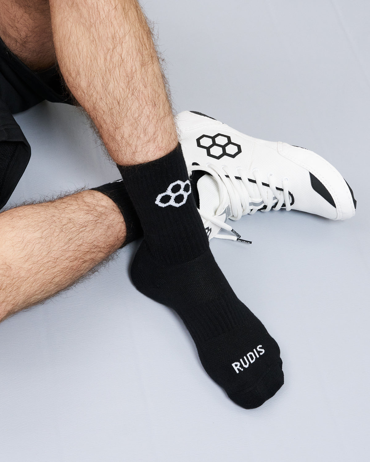 Person wearing black socks with a logo and 'RUDIS' text, paired with a white sneaker on a light gray background.
