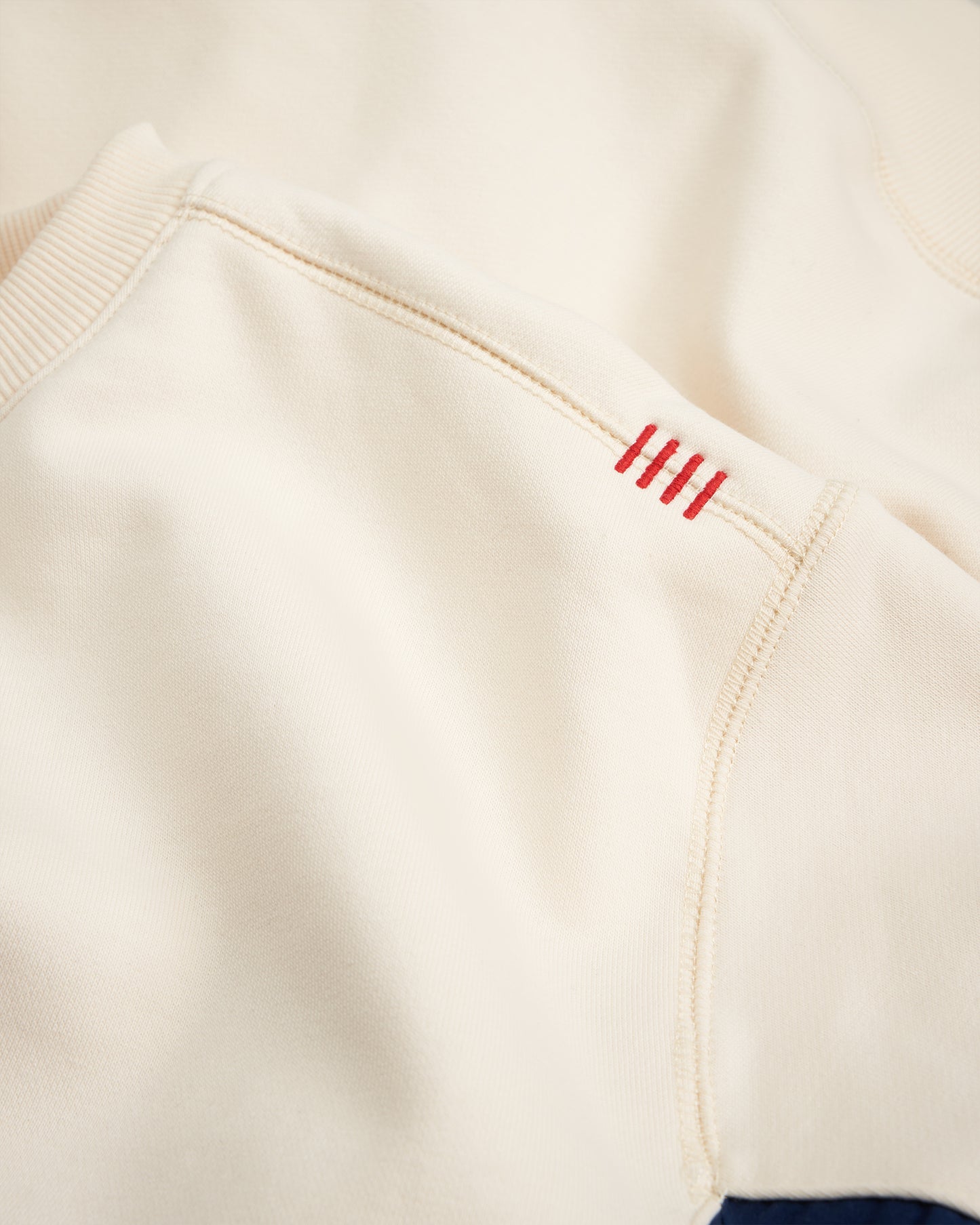A close-up view of a cream-colored sweatshirt showing intricate stitching details and a vibrant red decorative element on the sleeve