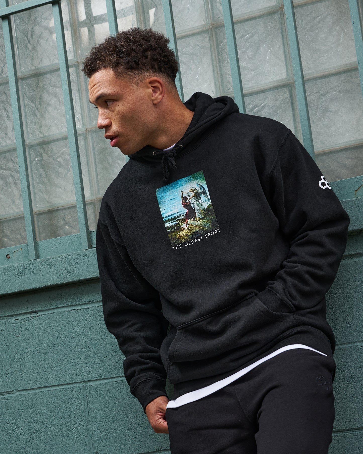 Person wearing a black hoodie with a graphic design, standing against a teal wall.