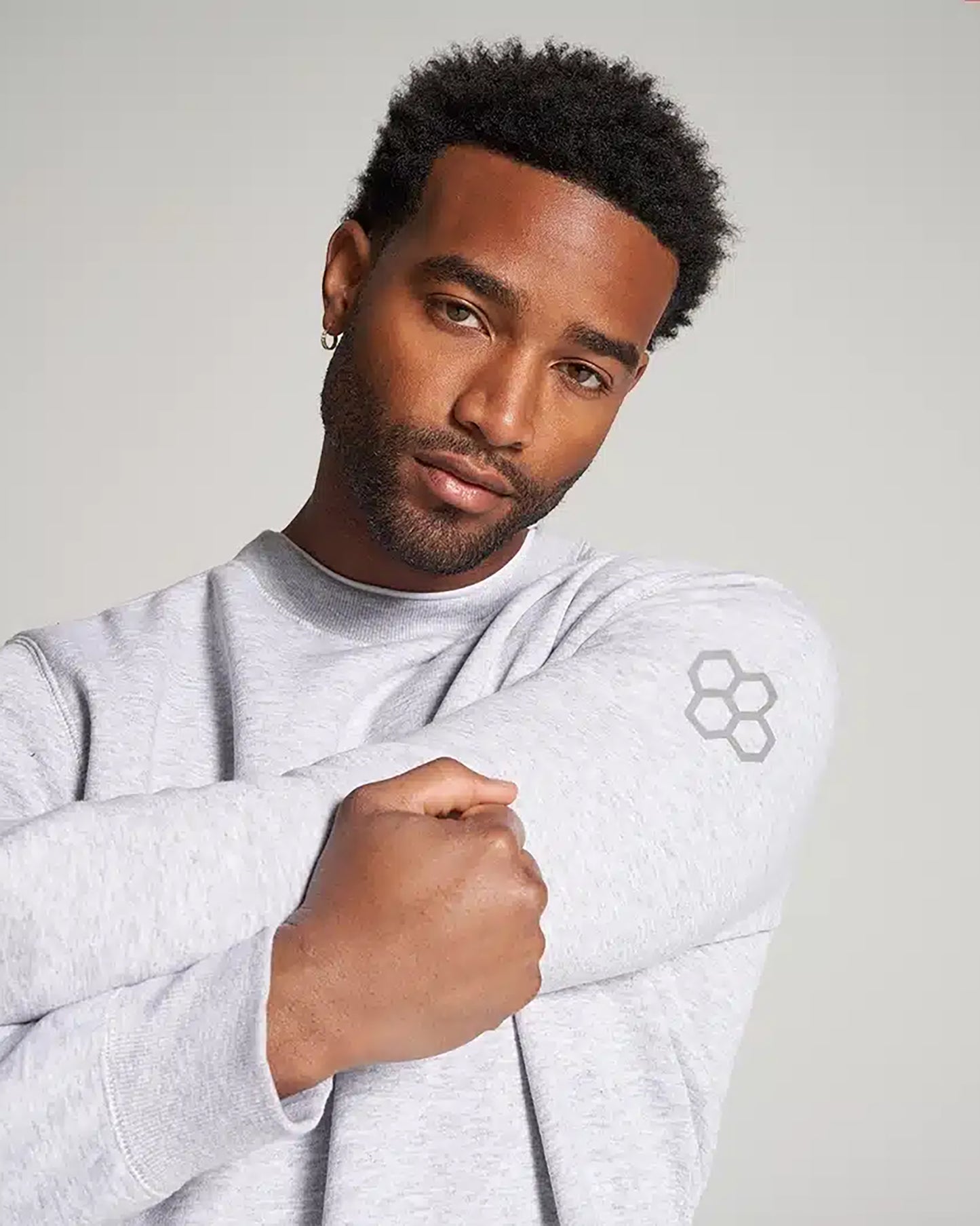 A stylish man poses in a light gray sweatshirt showcasing a subtle hexagonal logo on his arm while he gently crosses his arms
