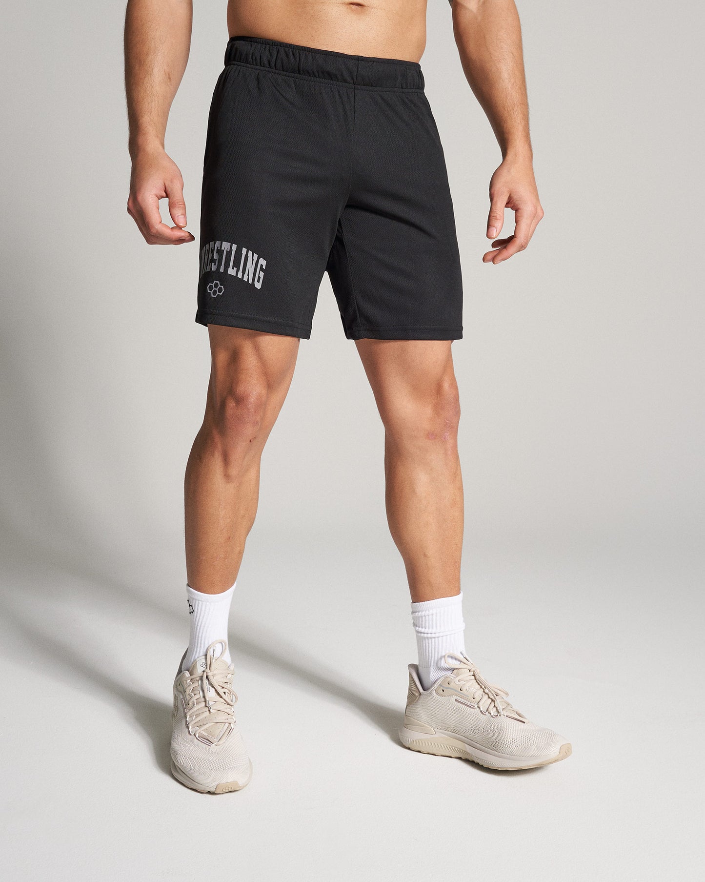 A male model stands confidently in athletic black shorts labeled WRESTLING and neutral-toned shoes showcasing an active lifestyle