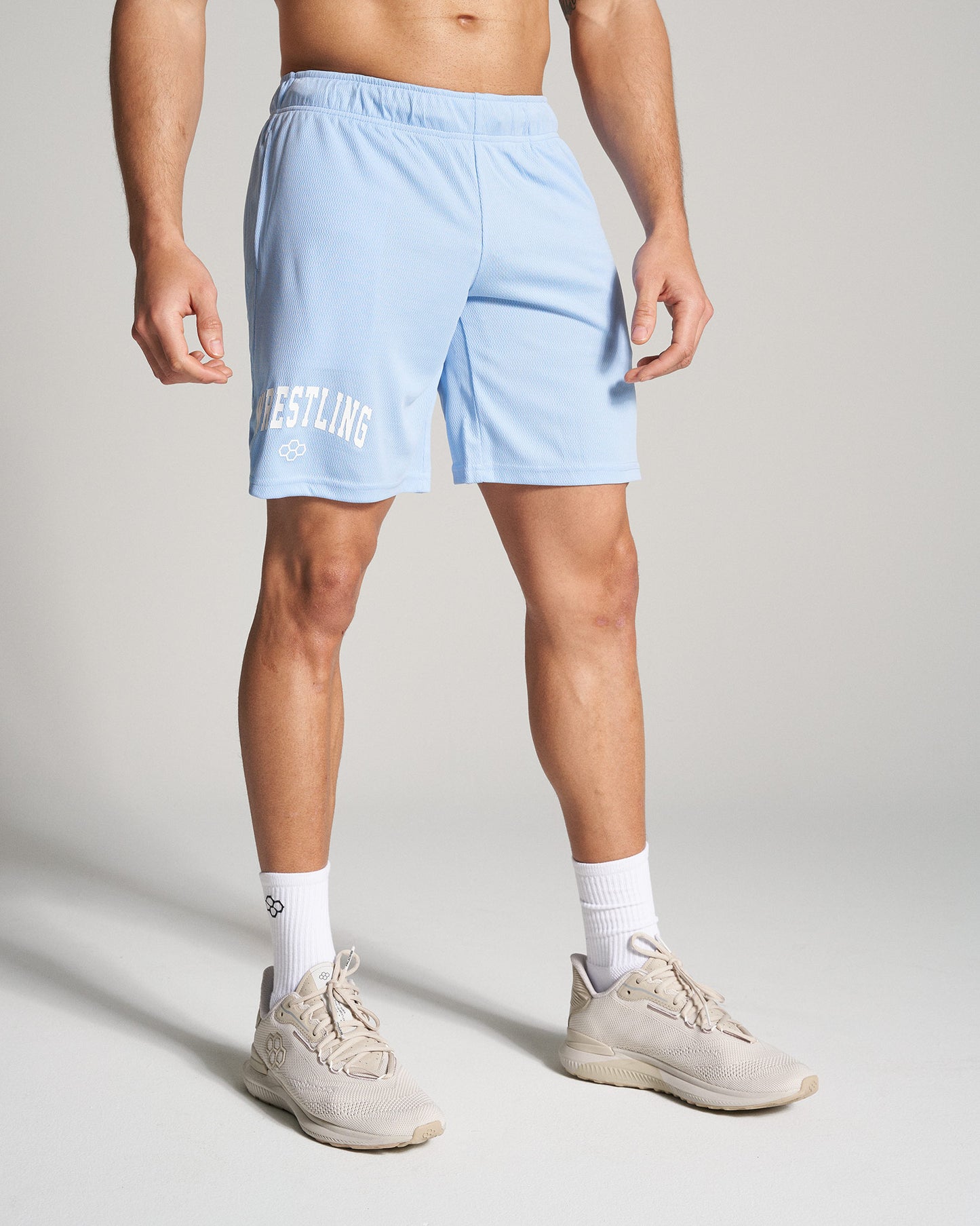 A muscular man is wearing light blue athletic shorts with WRESTLING printed on the side paired with beige running shoes and white socks standing against a neutral background
