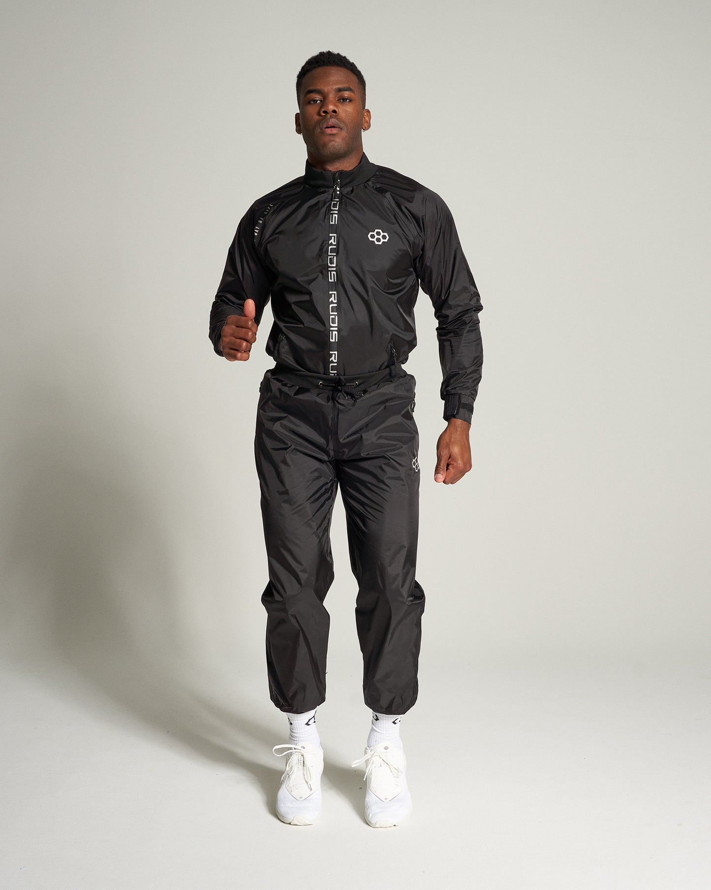 A male model is showcasing a stylish black athletic tracksuit complete with matching pants set against a light background