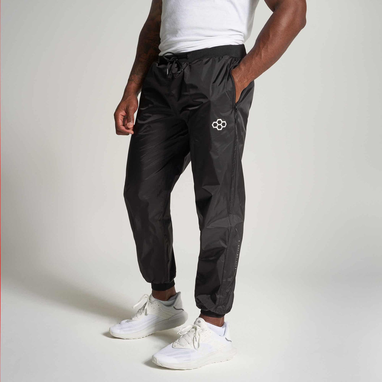 A male model wearing black track pants and white sneakers posing in a relaxed manner against a neutral background