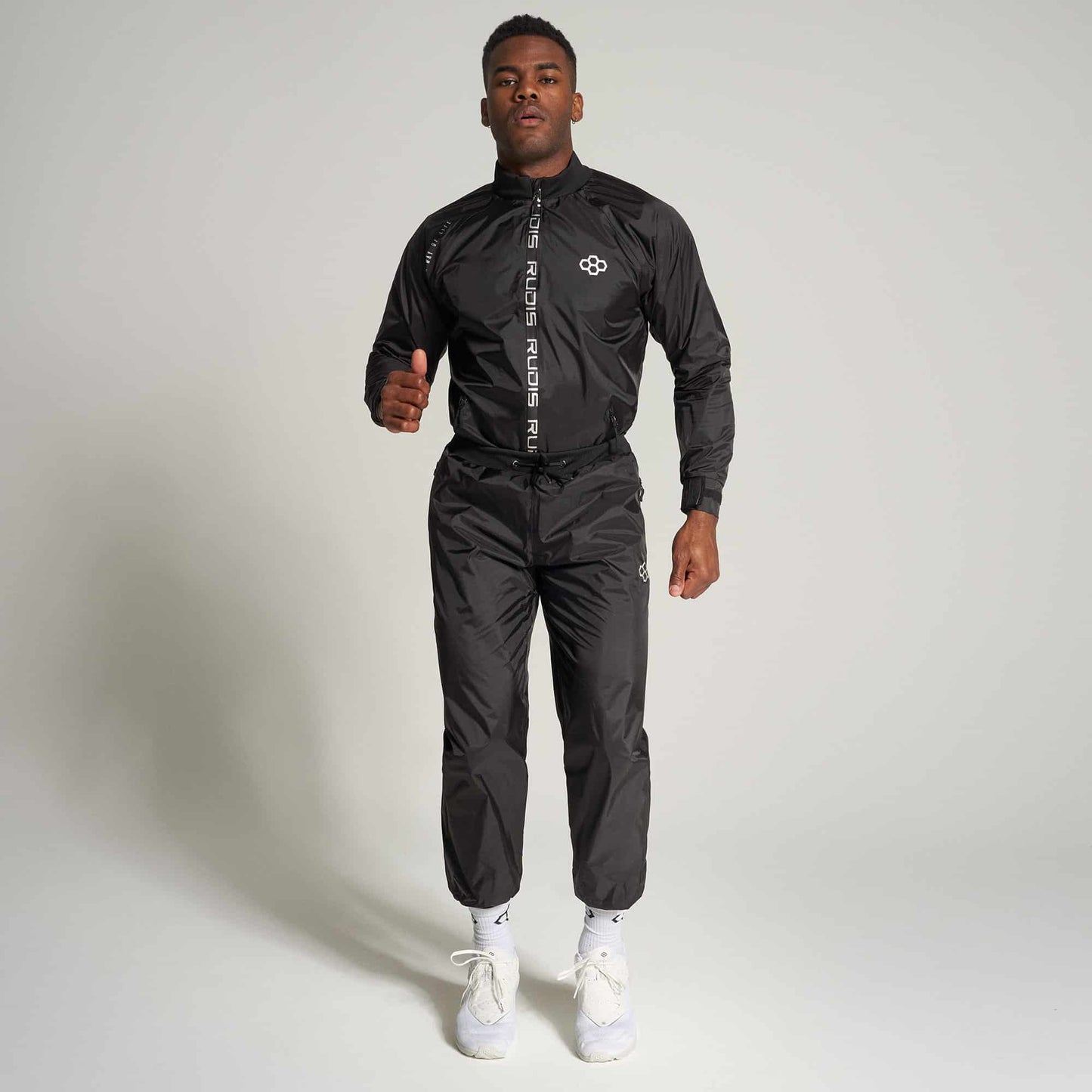 A male model stands confidently in a sleek black athletic outfit featuring a zip-up jacket and matching pants alongside white sneakers against a neutral background