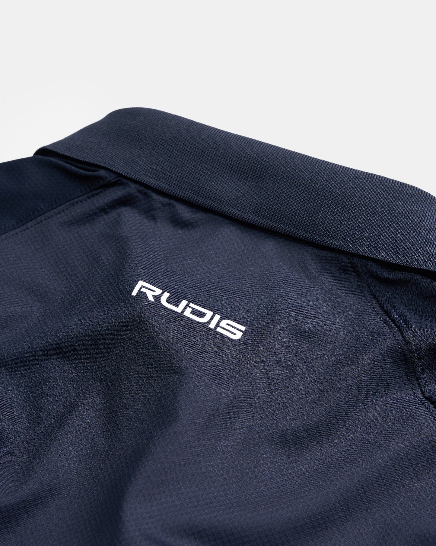A close-up view of a navy blue sports polo featuring a textured collar and the RUDIS logo embroidered in white