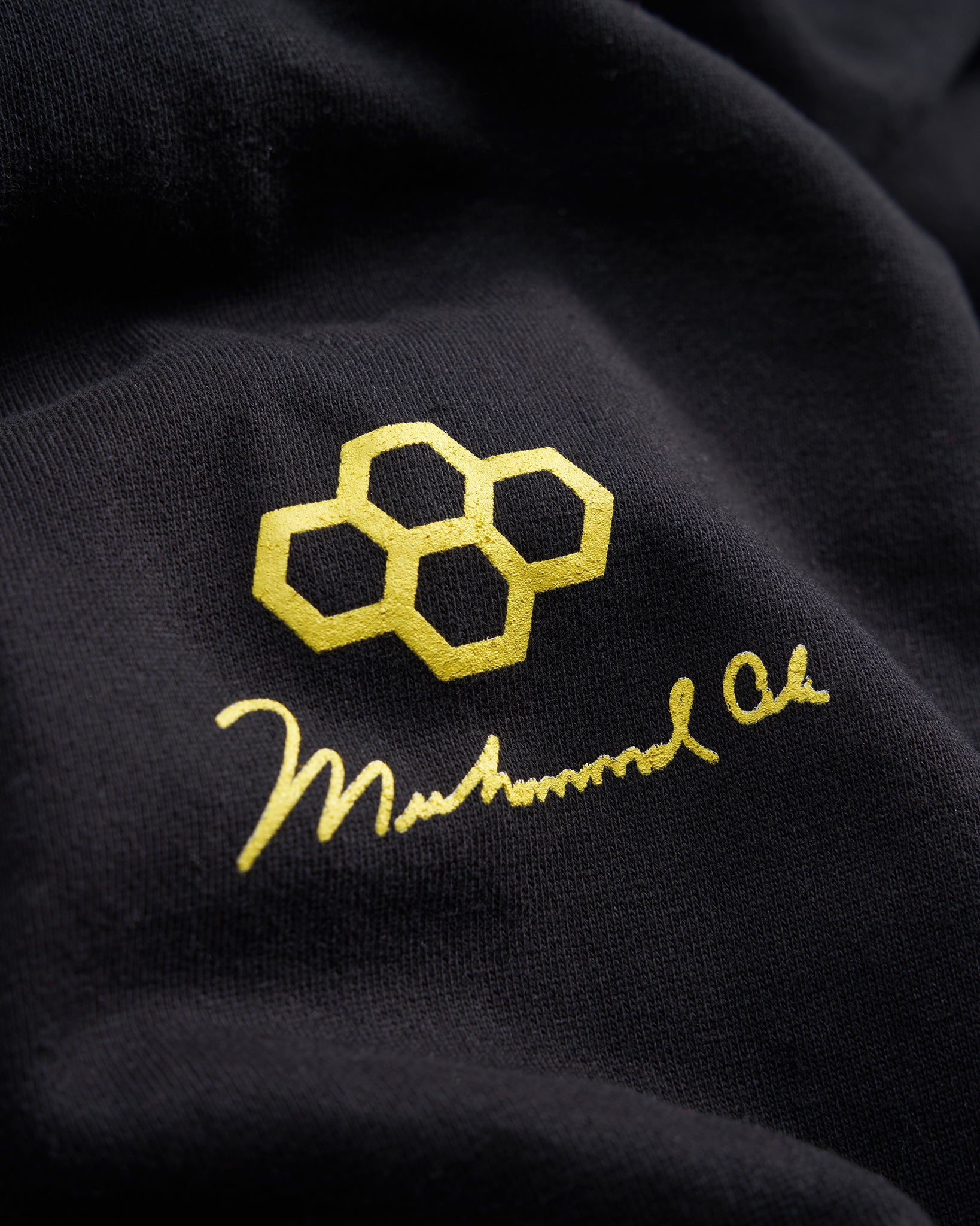 A close-up view of a black fabric showcasing a golden logo featuring interconnected hexagons and the signature of Muhammad Ali